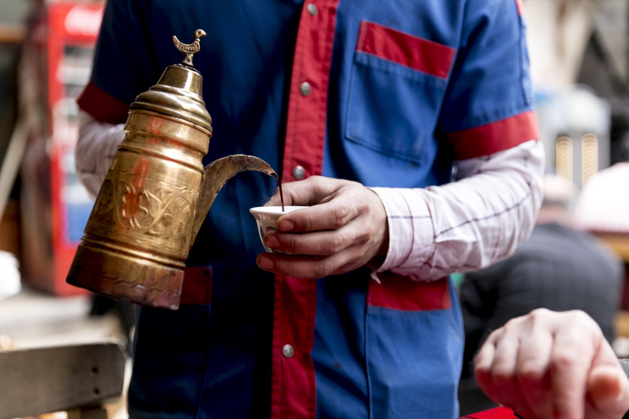 A person in a blue and red uniform pours coffee from an ornate brass pot into a small cup, with another’s arm visible—an image captured by Seattles best commercial food lifestyle photographer in an outdoor market setting. captured by seattle food and lifestyle photographer Brooke Fitts