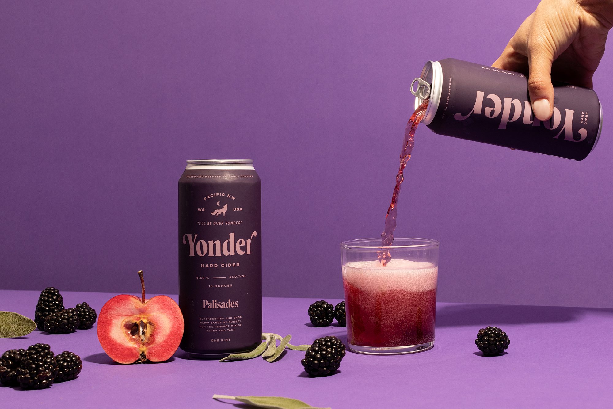 Seattle's best commercial food and hospitality photographer A hand pours Yonder Hard Cider from a can into a glass, surrounded by blackberries, a halved apple, sage leaves, and another can—all artfully styled by Seattles best commercial food lifestyle photographer against a purple background. captured by seattle's best food and lifestyle photographer Brooke Fitts