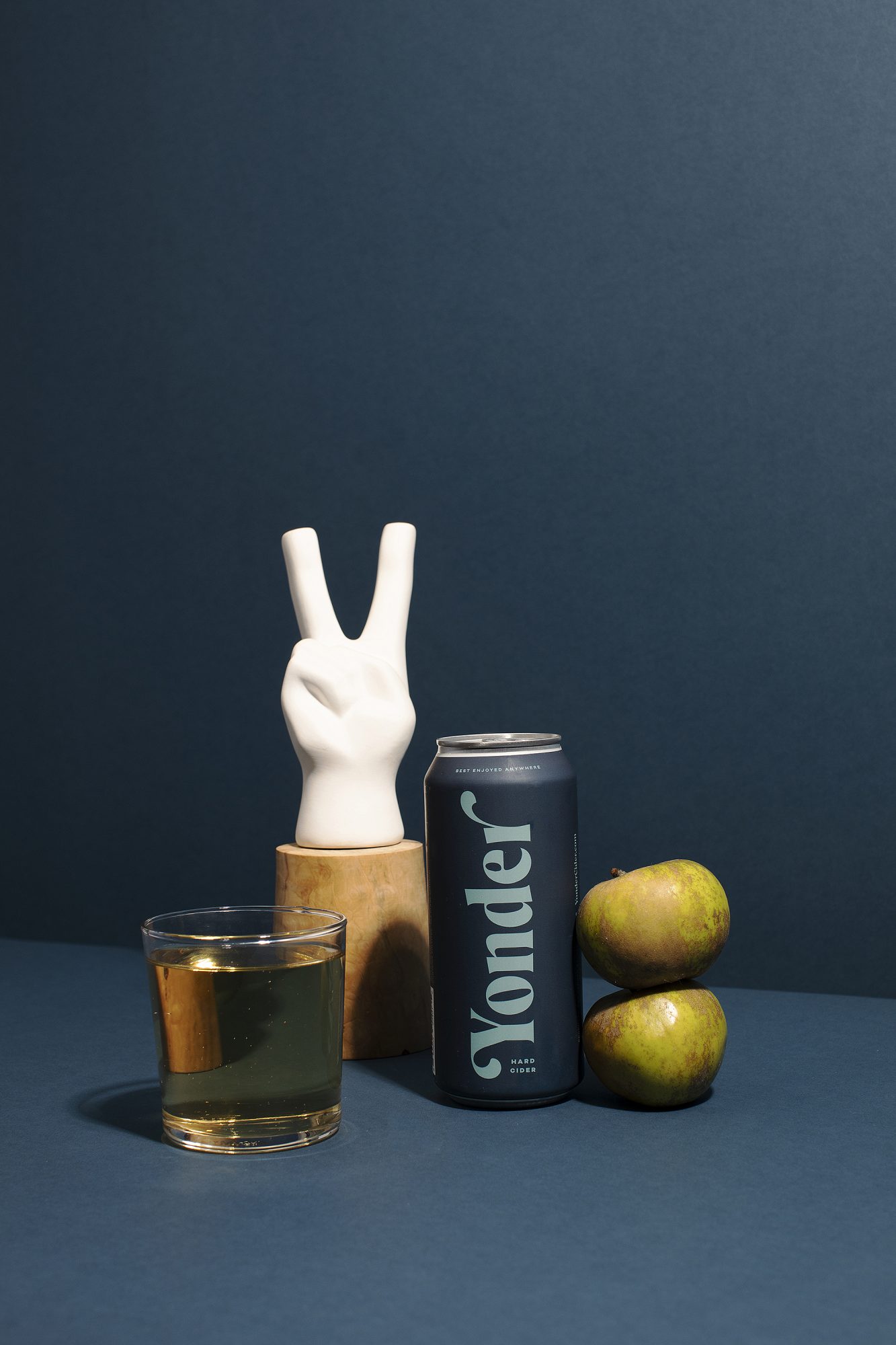 A can labeled Yonder stands next to two green apples, a glass of juice, and a white ceramic hand making a peace sign—an artful scene by Seattles best commercial food lifestyle photographer, set against a dark blue background. captured by seattle food and lifestyle photographer Brooke Fitts