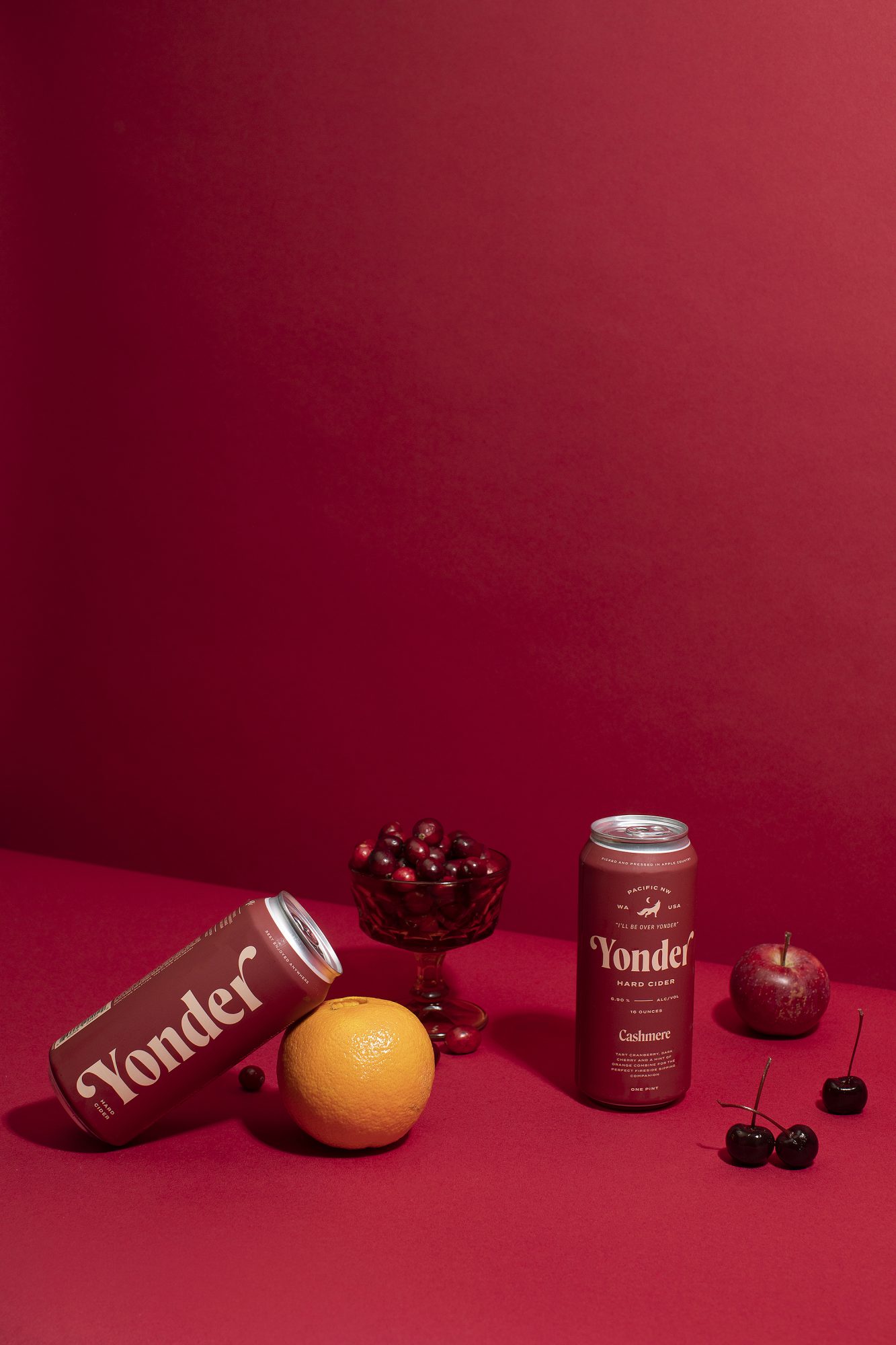 Two Yonder cans, one upright and one lying down, are on a red surface with an orange, an apple, cherries, and a bowl of cherries—all artfully captured by Seattles best commercial food lifestyle photographer. captured by seattle food and lifestyle photographer Brooke Fitts
