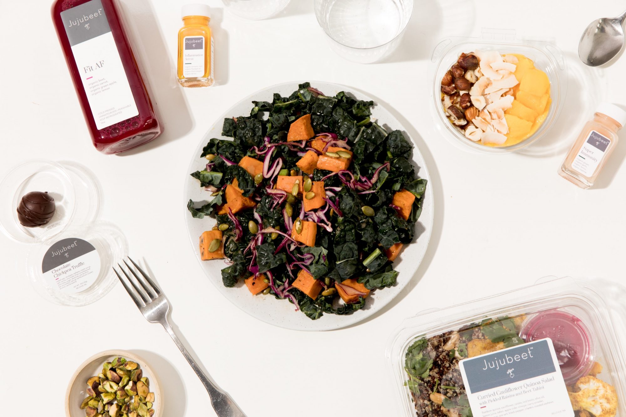 Seattle's best commercial food and hospitality photographer A plate of kale salad with sweet potatoes and red cabbage sits on a white table, styled by Seattles best commercial food lifestyle photographer, surrounded by a red juice bottle, nut containers, a fork, and other packaged food items. captured by seattle's best food and lifestyle photographer Brooke Fitts