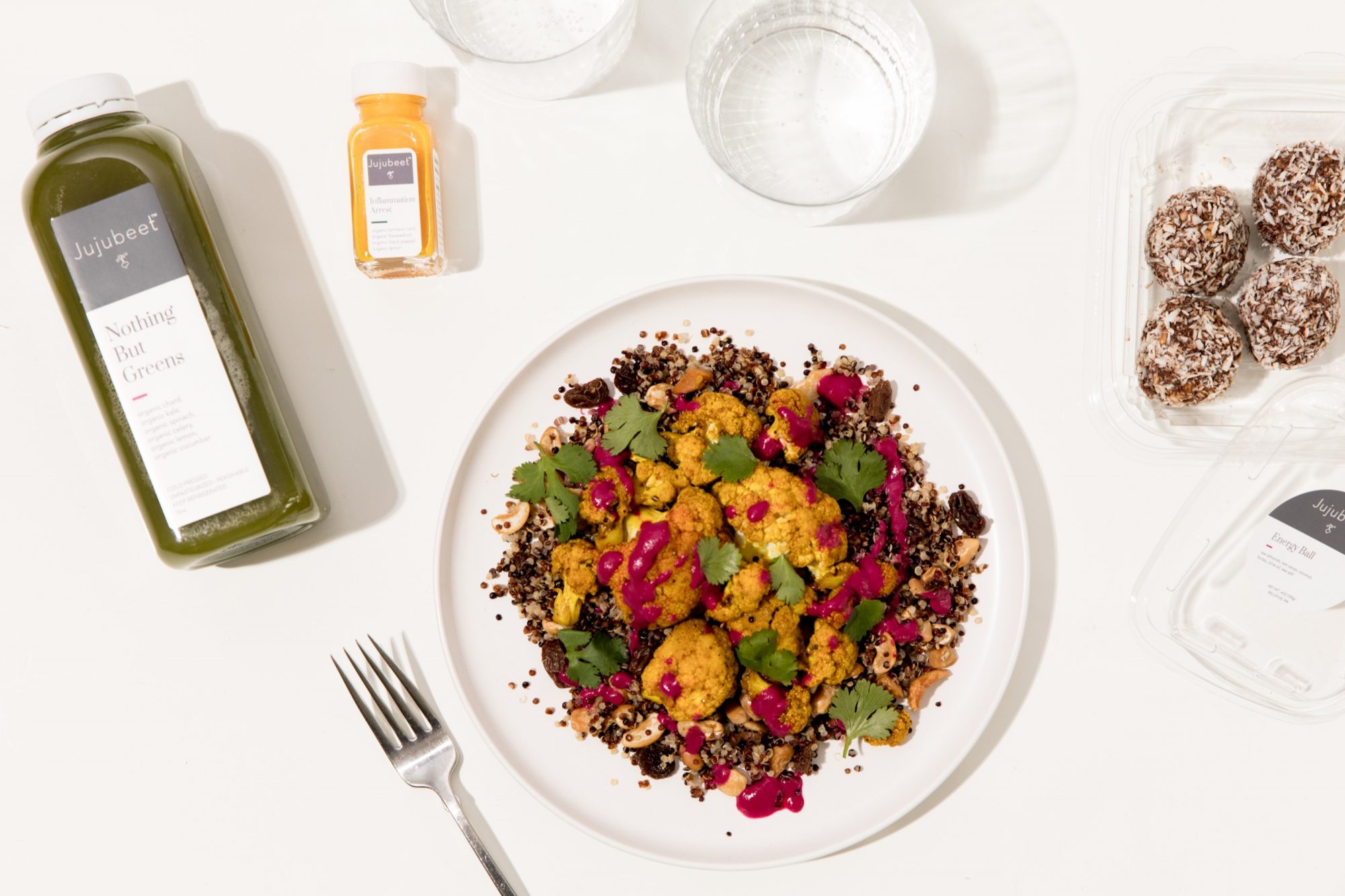 Seattle's best commercial food and hospitality photographer A plate of quinoa salad with roasted cauliflower, fresh cilantro, and bright pink sauce is artfully arranged by Seattles best commercial food lifestyle photographer, surrounded by green juice, a turmeric shot, water glasses, a fork, and energy bites on a white surface. captured by seattle's best food and lifestyle photographer Brooke Fitts