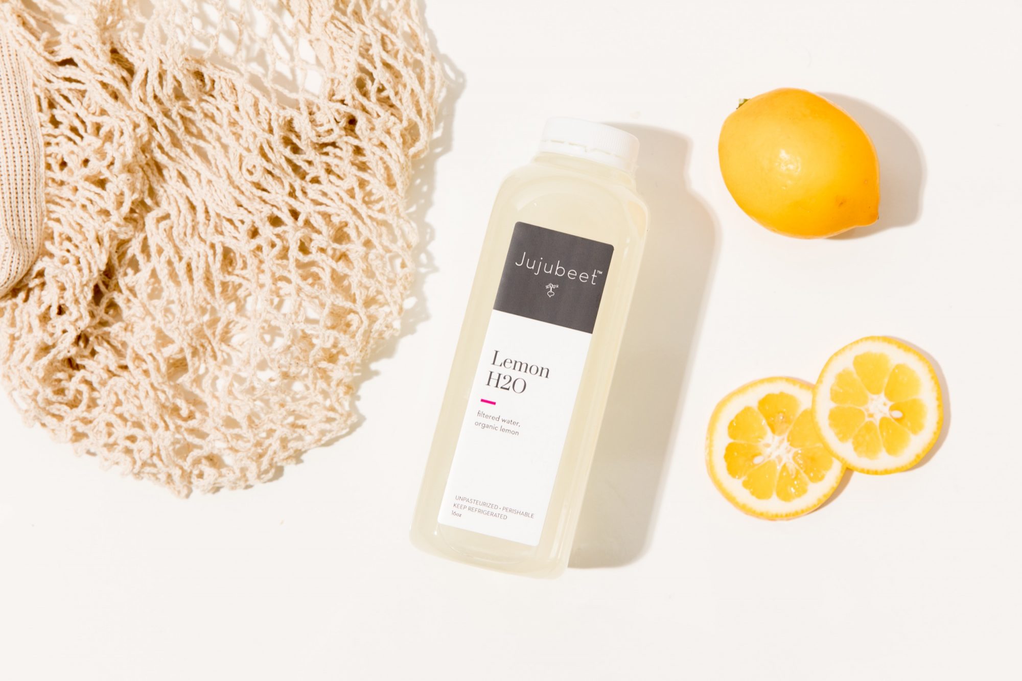 Seattle's best commercial food and hospitality photographer A clear bottle labeled Jujubeet Lemon H2O lies on a white surface next to a cut lemon, a whole lemon, and a beige mesh bag, artfully captured by Seattles best commercial food lifestyle photographer. captured by seattle's best food and lifestyle photographer Brooke Fitts