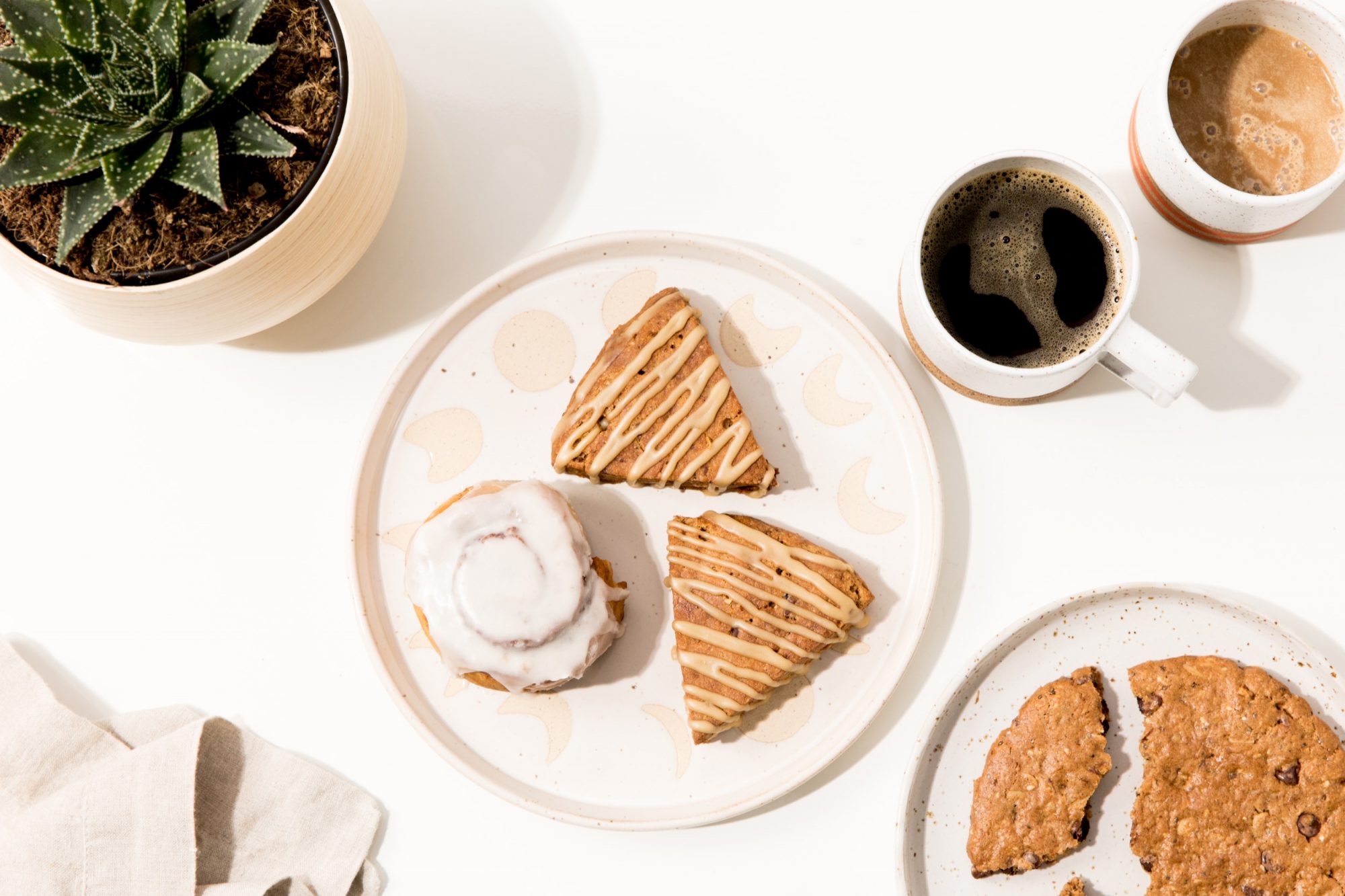 Seattle's best commercial food and hospitality photographer A plate with two glazed scones and an iced cinnamon roll sits on a white surface next to a cookie, a cup of coffee, and a potted succulent plant—captured by Seattles best commercial food lifestyle photographer in bright, natural lighting. captured by seattle's best food and lifestyle photographer Brooke Fitts