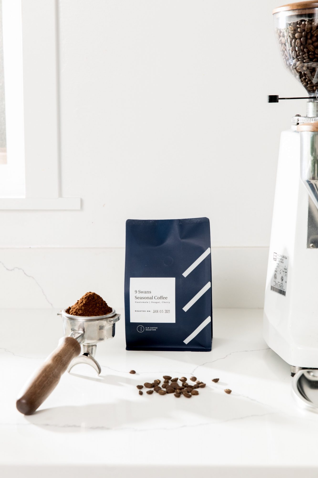 A navy blue coffee bag stands on a white countertop next to a portafilter filled with ground coffee, scattered coffee beans, and a coffee grinder—capturing the craft through Seattles best commercial food lifestyle photographer. captured by seattle food and lifestyle photographer Brooke Fitts