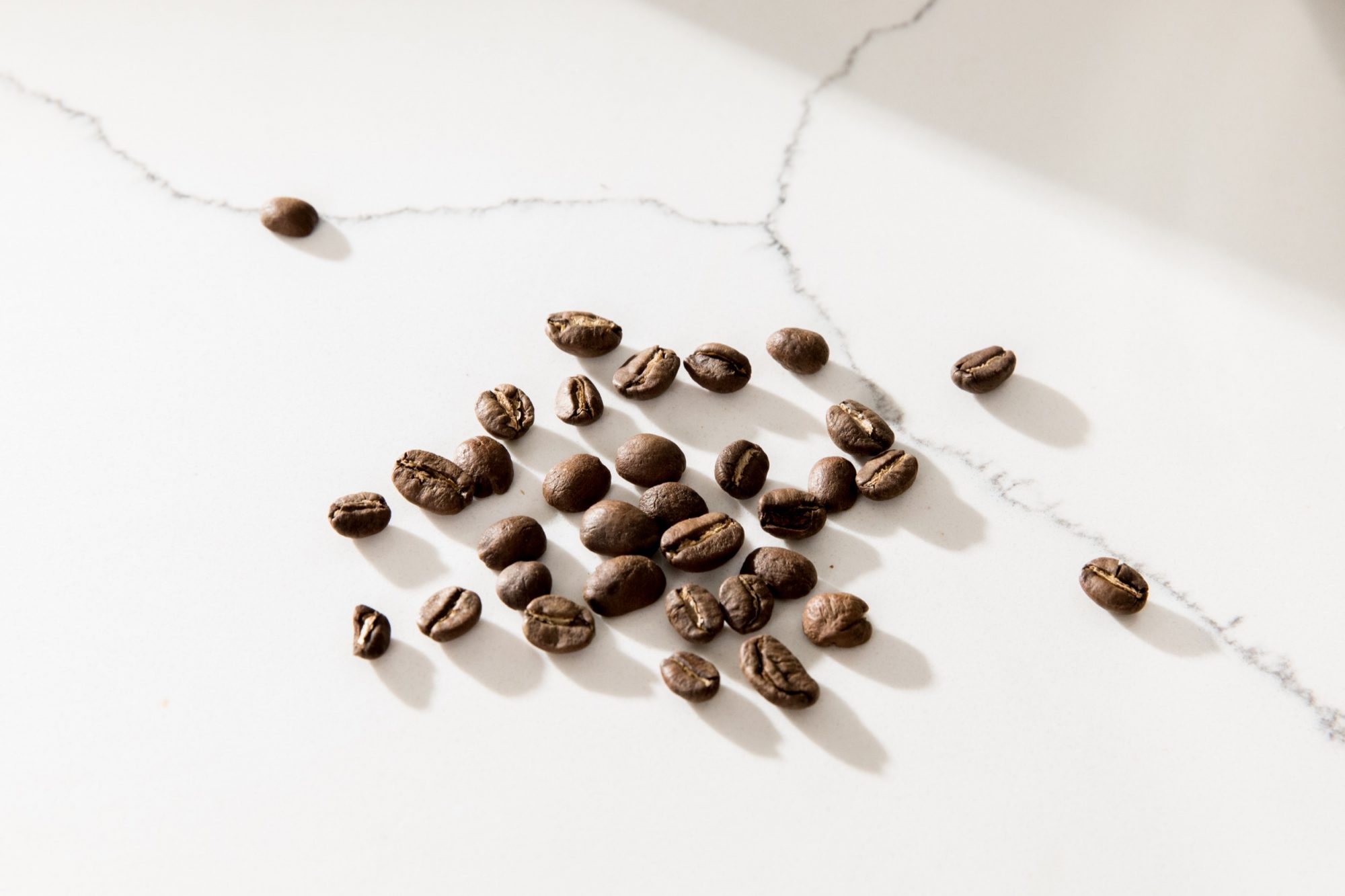Scattered coffee beans on a white marble surface with visible grey veins, illuminated by natural light casting soft shadows, captured by Seattles best commercial food lifestyle photographer. captured by seattle food and lifestyle photographer Brooke Fitts