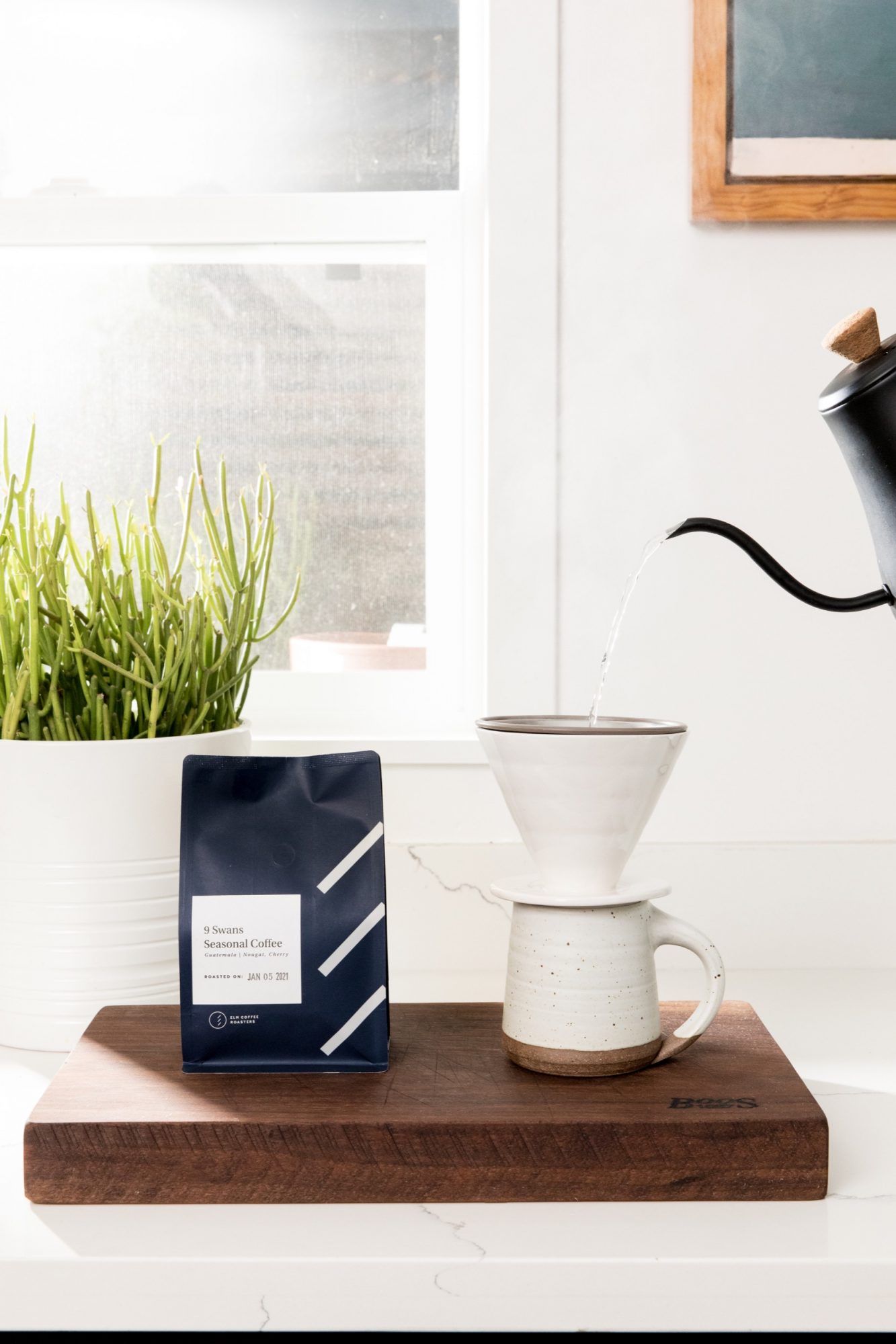 A kettle pours hot water into a white ceramic pour-over coffee dripper set on a matching mug. Captured by Seattles best commercial food lifestyle photographer, the cozy scene includes a coffee bag, wooden board, and a green plant by a sunny window. captured by seattle food and lifestyle photographer Brooke Fitts