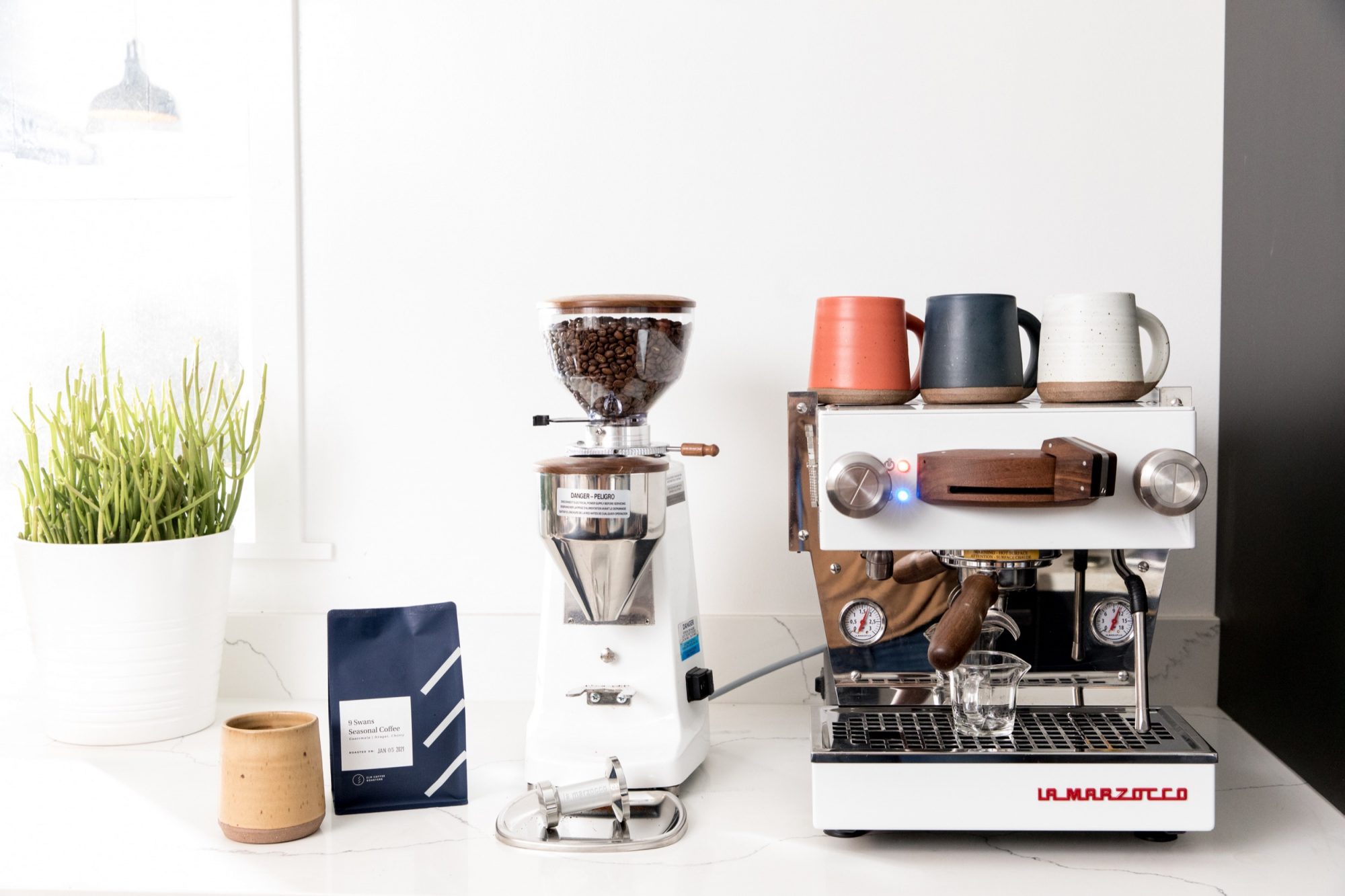A white espresso machine and coffee grinder sit on a countertop with mugs on top. Captured by Seattles best commercial food lifestyle photographer, this bright setup is complete with a potted plant, coffee beans, and a ceramic cup. captured by seattle food and lifestyle photographer Brooke Fitts