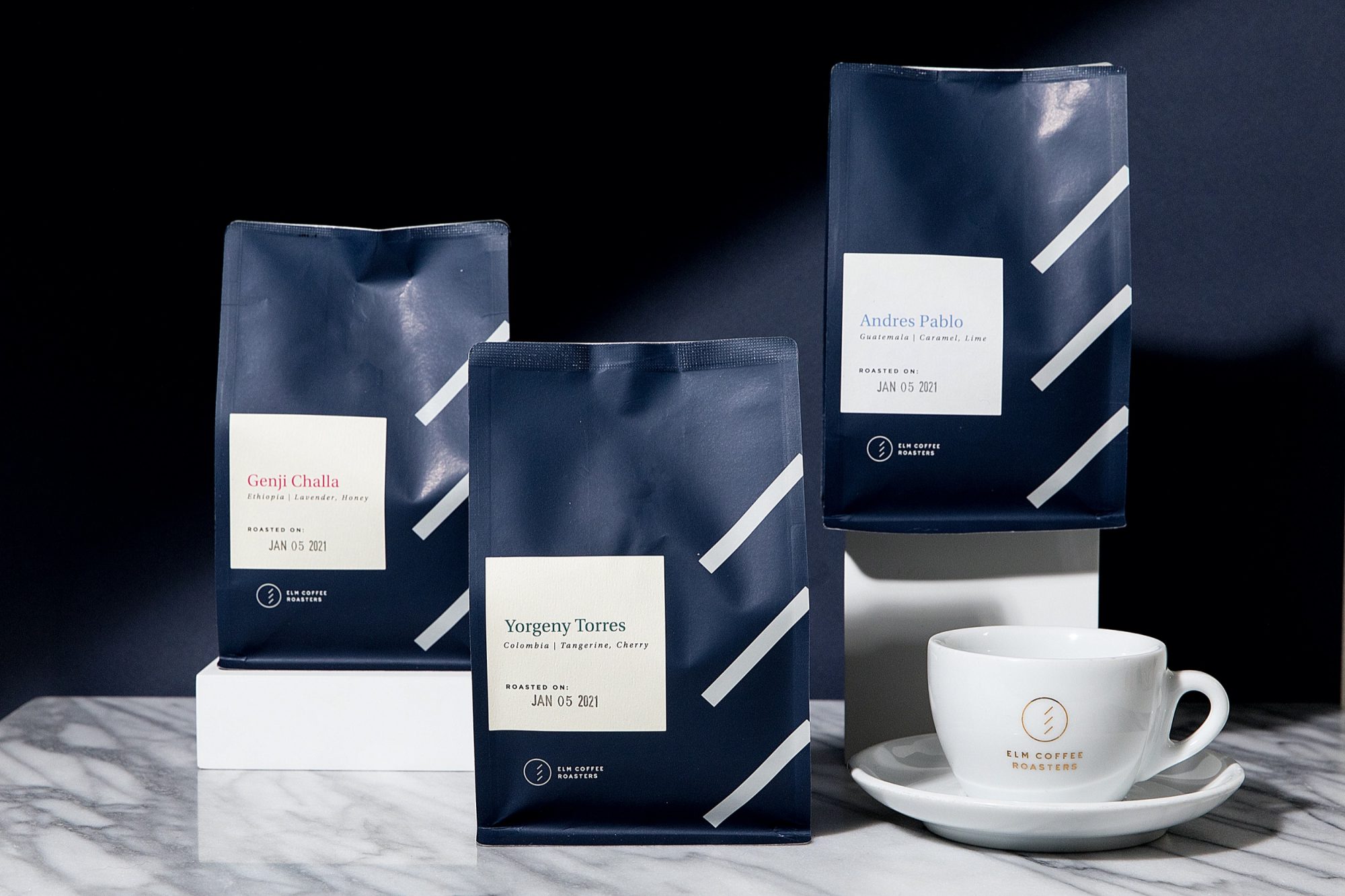 Three dark blue coffee bags with white labels and diagonal stripes are displayed on a marble surface next to a white coffee cup—styled by Seattles best commercial food lifestyle photographer. Each bag features different producers and roast dates. captured by seattle food and lifestyle photographer Brooke Fitts