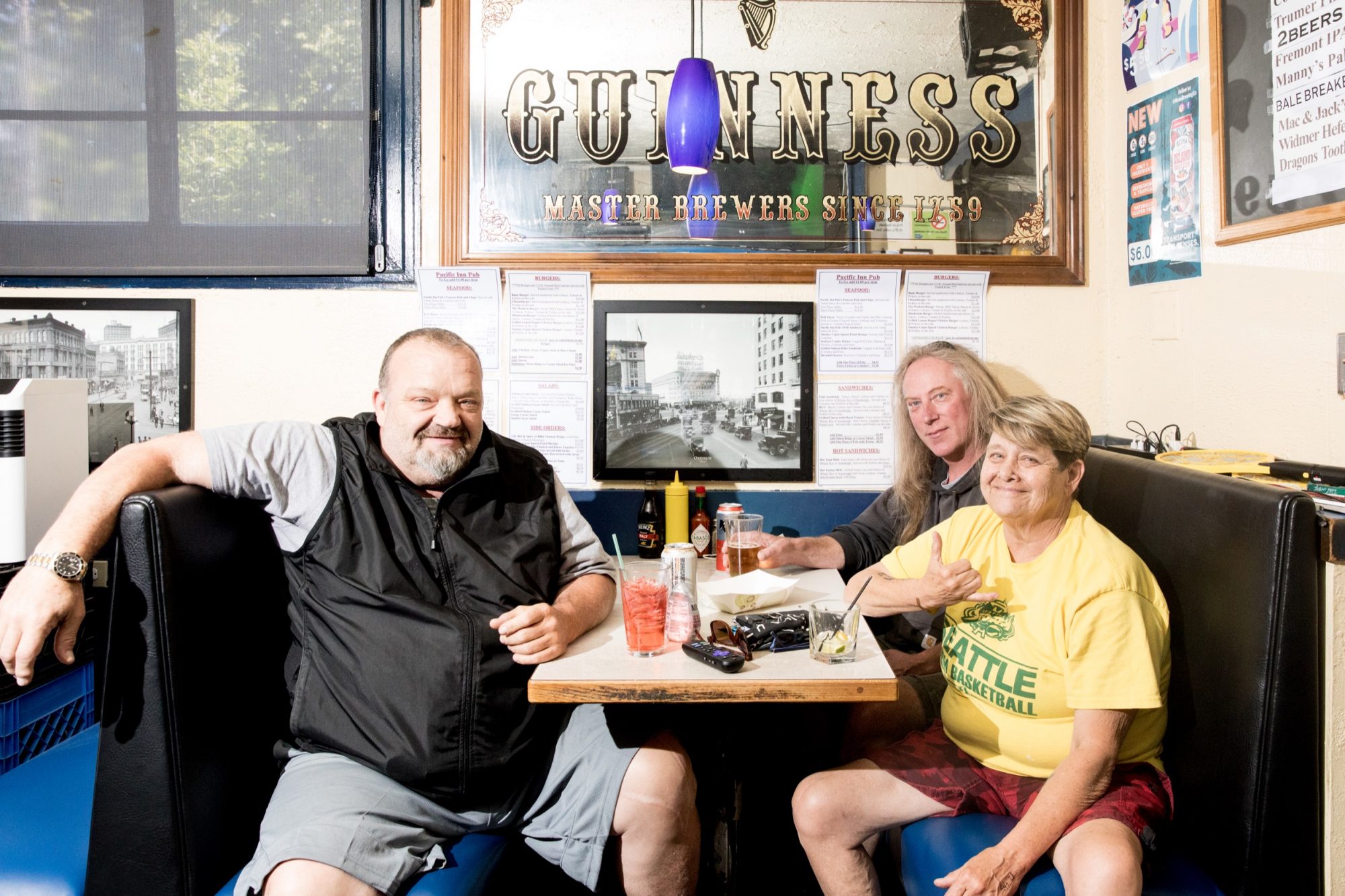 Seattle's best commercial food and hospitality photographer Three people sit in a restaurant booth with drinks and food on the table, smiling at the camera. Captured by Seattles best commercial food lifestyle photographer, the casual setting is brightly lit, with a Guinness sign and framed photos behind them. captured by seattle's best food and lifestyle photographer Brooke Fitts