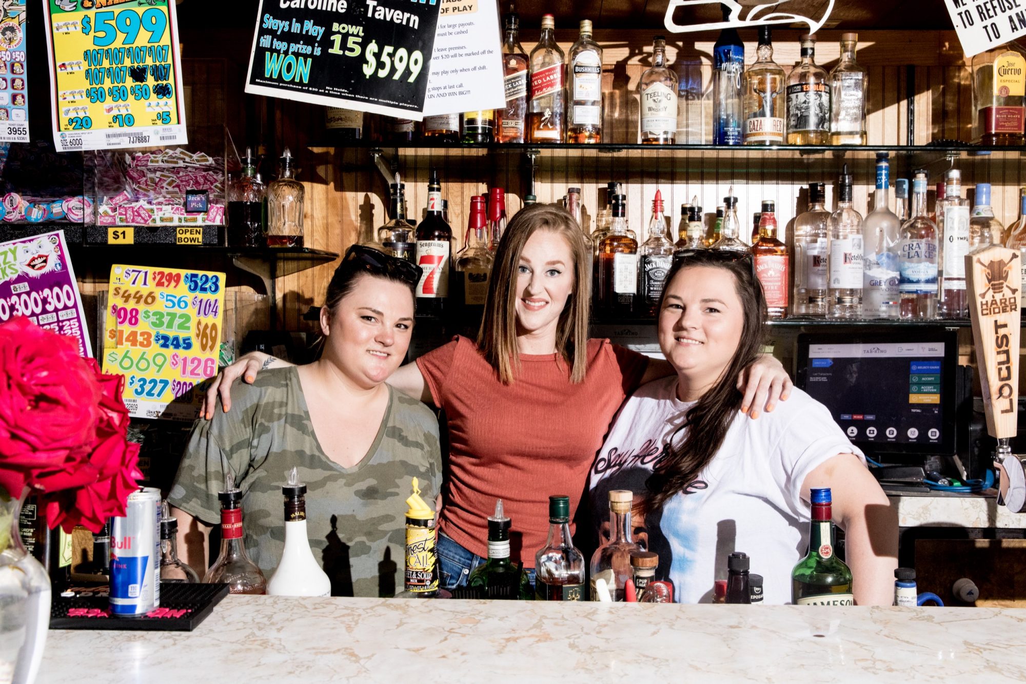 Seattle's best commercial food and hospitality photographer Three women stand behind a bar counter with various bottles of alcohol, smiling at the camera. Captured by Seattles best commercial food lifestyle photographer, theyre surrounded by colorful signs showcasing drink specials and prices. captured by seattle's best food and lifestyle photographer Brooke Fitts