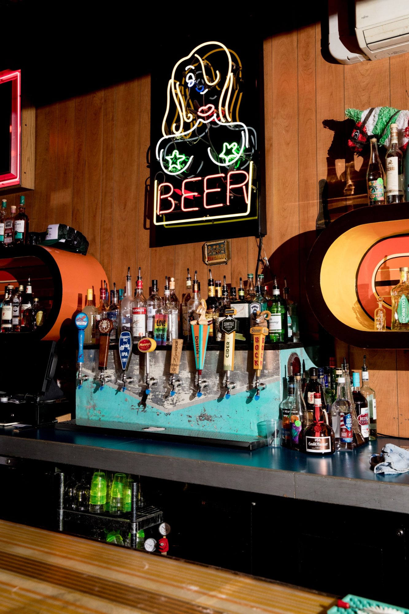 Seattle's best commercial food and hospitality photographer A bar counter with various liquor bottles and beer taps below a neon sign of a stylized woman and BEER in yellow lettering, captured by Seattles best commercial food lifestyle photographer. Wood-paneled walls and eclectic decor complete the scene. captured by seattle's best food and lifestyle photographer Brooke Fitts
