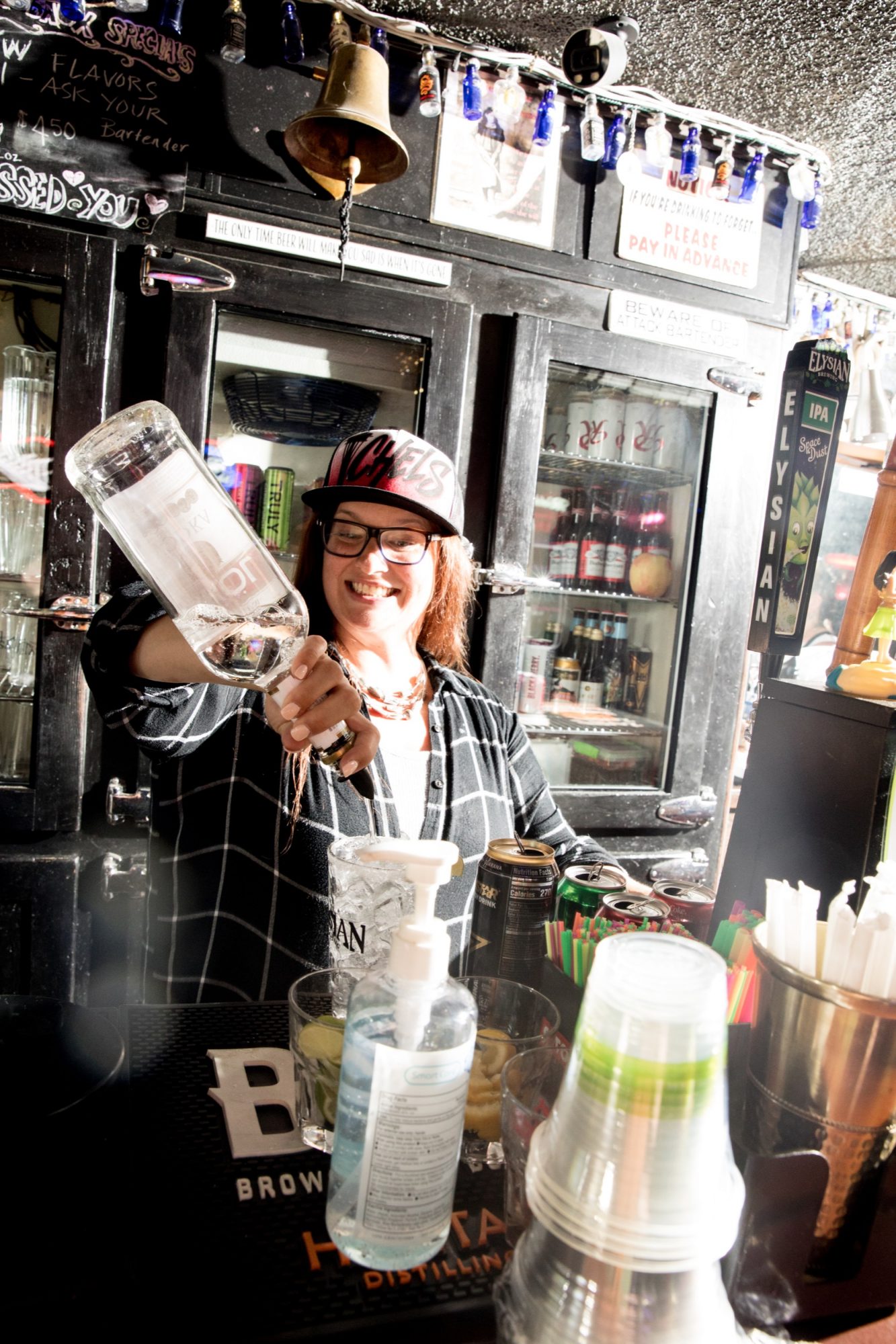 Seattle's best commercial food and hospitality photographer A smiling bartender in a plaid shirt and hat pours vodka behind a bar, surrounded by bottles and cocktail supplies, as bright light shines across the scene—captured by Seattles best commercial food lifestyle photographer. captured by seattle's best food and lifestyle photographer Brooke Fitts