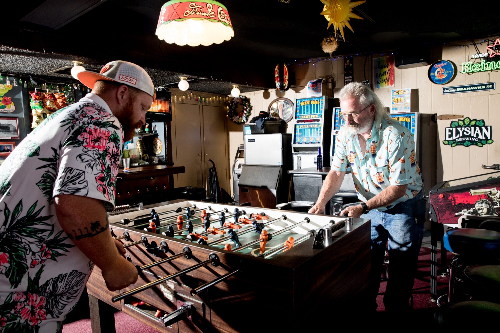 Seattle's best commercial food and hospitality photographer Two men play foosball in a dimly lit bar filled with retro signs and arcade machines, captured by Seattles best commercial food lifestyle photographer. One sports a floral shirt and cap; the other, a white beard, glasses, and light-blue shirt. captured by seattle's best food and lifestyle photographer Brooke Fitts