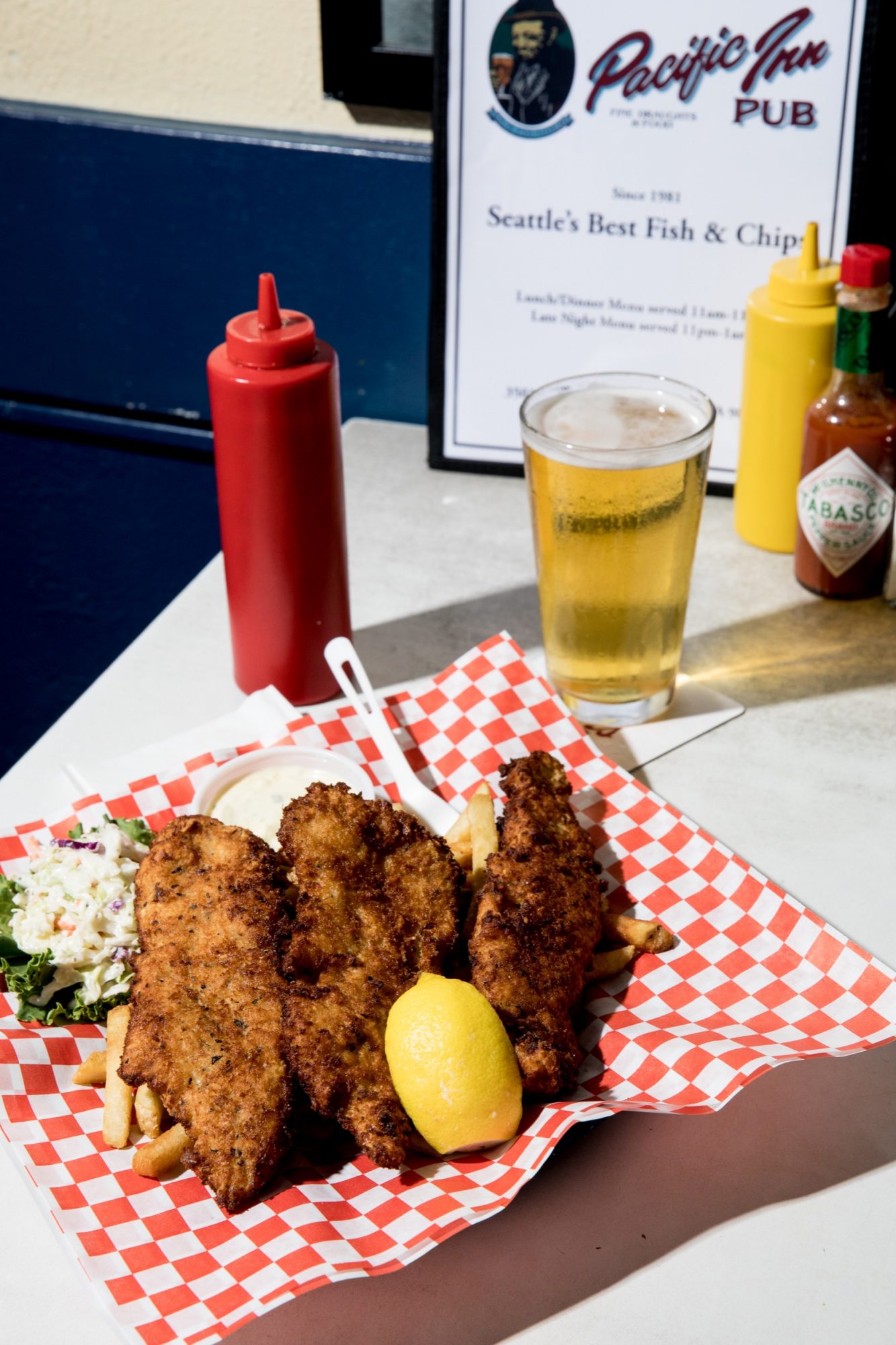 Seattle's best commercial food and hospitality photographer A basket of fried fish, fries, and a lemon wedge sits on red checkered paper next to coleslaw, perfectly captured by Seattles best commercial food lifestyle photographer with a glass of beer and Pacific Inn Pub menu in the background. captured by seattle's best food and lifestyle photographer Brooke Fitts