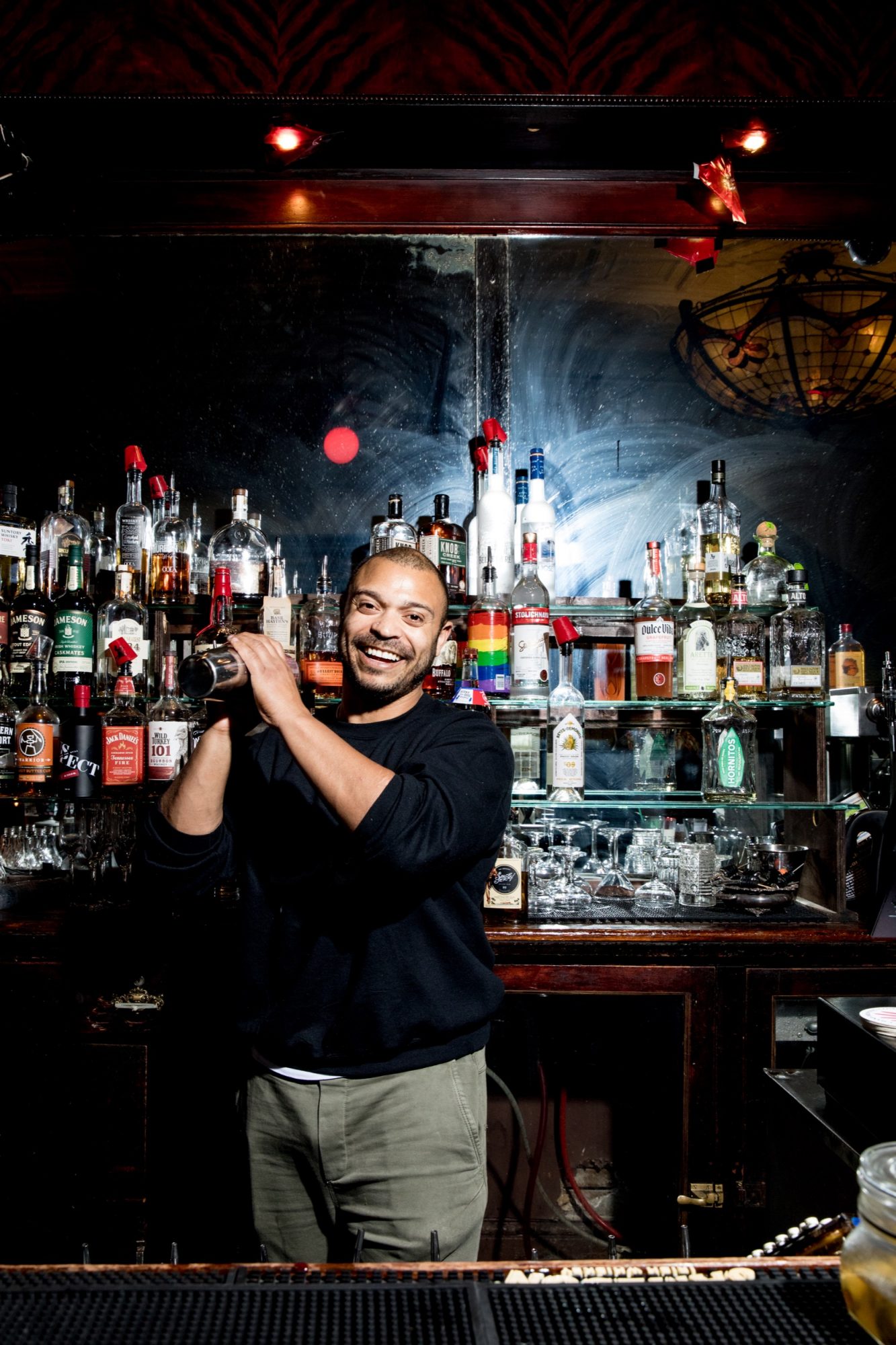 Seattle's best commercial food and hospitality photographer A smiling bartender stands behind a bar, shaking a cocktail shaker. Captured by Seattles best commercial food lifestyle photographer, shelves filled with various bottles of liquor and glassware are visible in the background. captured by seattle's best food and lifestyle photographer Brooke Fitts