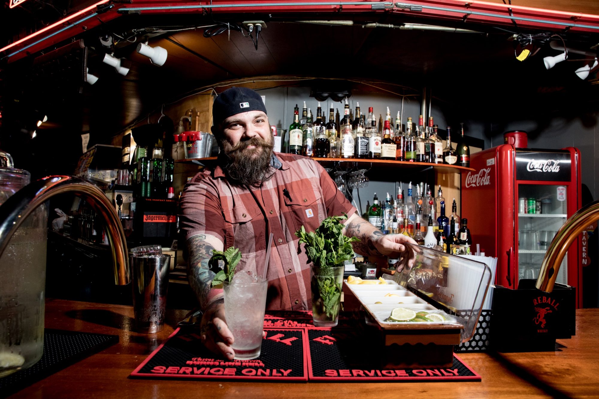 Seattle's best commercial food and hospitality photographer A bearded bartender in a plaid shirt and backwards cap smiles while preparing cocktails at a bar, captured by Seattles best commercial food lifestyle photographer, with bottles, garnishes, and a Coca-Cola fridge in the background. captured by seattle's best food and lifestyle photographer Brooke Fitts