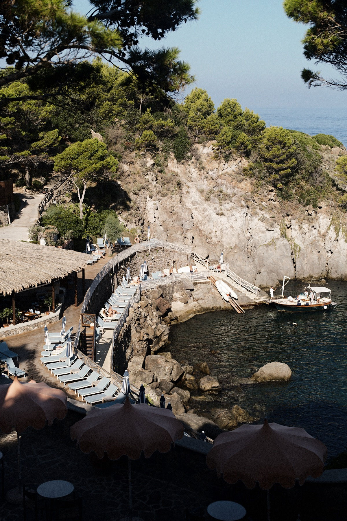 Seattle's best commercial food and hospitality photographer A rocky coastal area with sun loungers, umbrellas, and a wooden boardwalk by the sea, captured by Seattles best commercial food lifestyle photographer. Lush green trees and cliffs surround a small boat docked near the water’s edge. captured by seattle's best food and lifestyle photographer Brooke Fitts