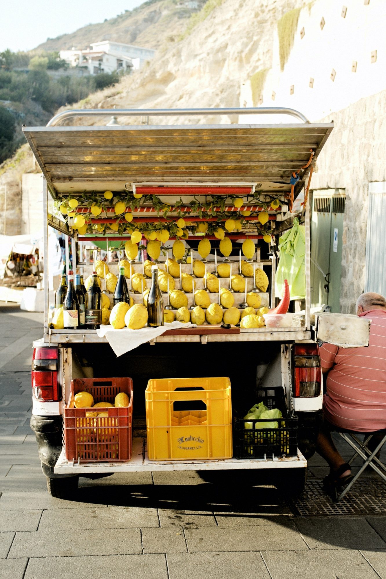 Seattle's best commercial food and hospitality photographer A small truck, styled by Seattles best commercial food lifestyle photographer, is decorated with lemons and bottles and displays crates of fruit and drinks for sale. Parked on a sunny street by a stone wall, a person sits to the right, partially out of view. captured by seattle's best food and lifestyle photographer Brooke Fitts