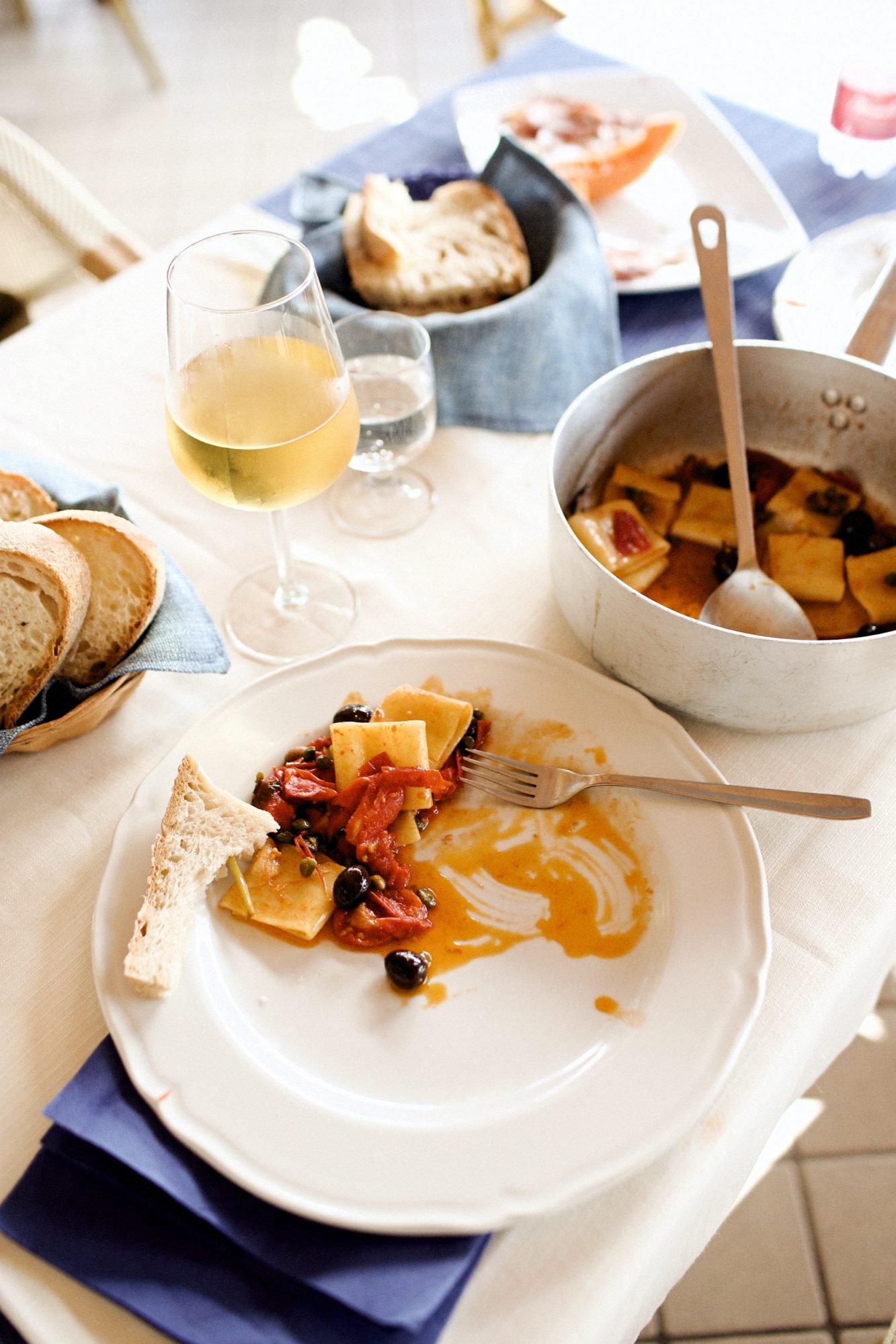Seattle's best commercial food and hospitality photographer A white plate with pasta, tomatoes, olives, and a slice of bread sits on a table styled by Seattles best commercial food lifestyle photographer, complete with white wine, a basket of sliced bread, and pasta on a cream tablecloth with a blue napkin. captured by seattle's best food and lifestyle photographer Brooke Fitts