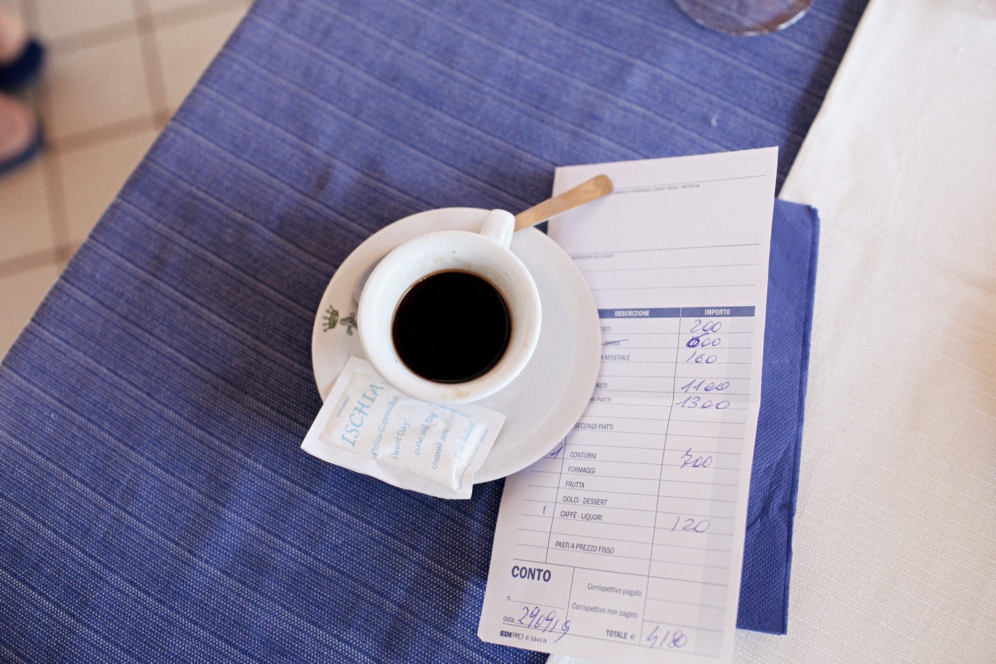 Seattle's best commercial food and hospitality photographer A cup of black coffee on a saucer with a sugar packet and spoon rests on a restaurant bill atop a blue tablecloth, captured by Seattles best commercial food lifestyle photographer. captured by seattle's best food and lifestyle photographer Brooke Fitts
