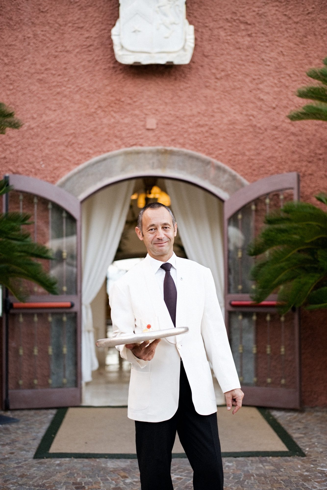 Seattle's best commercial food and hospitality photographer A waiter in a white jacket and dark tie stands outside an arched entrance, holding a tray with a drink—a scene expertly captured by Seattles best commercial food lifestyle photographer. Open doors, white curtains, and greenery complete the elegant backdrop. captured by seattle's best food and lifestyle photographer Brooke Fitts