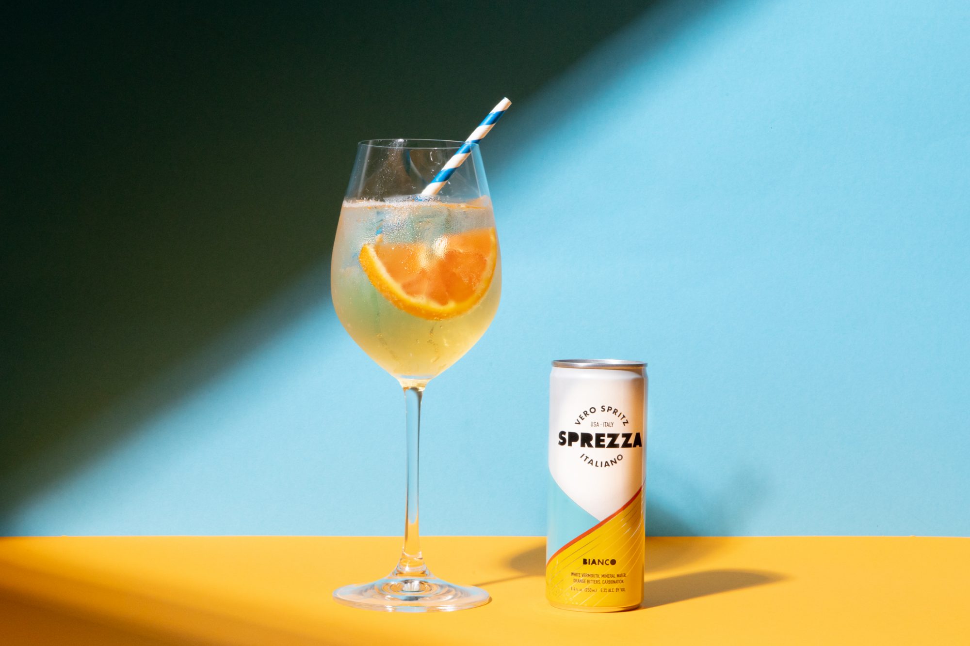 Seattle's best commercial food and hospitality photographer A wine glass with a striped straw and orange slices holds a sparkling drink. Next to it, a can labeled Sprezza Bianco rests on a yellow surface with a blue background—shot by Seattles best commercial food lifestyle photographer. captured by seattle's best food and lifestyle photographer Brooke Fitts