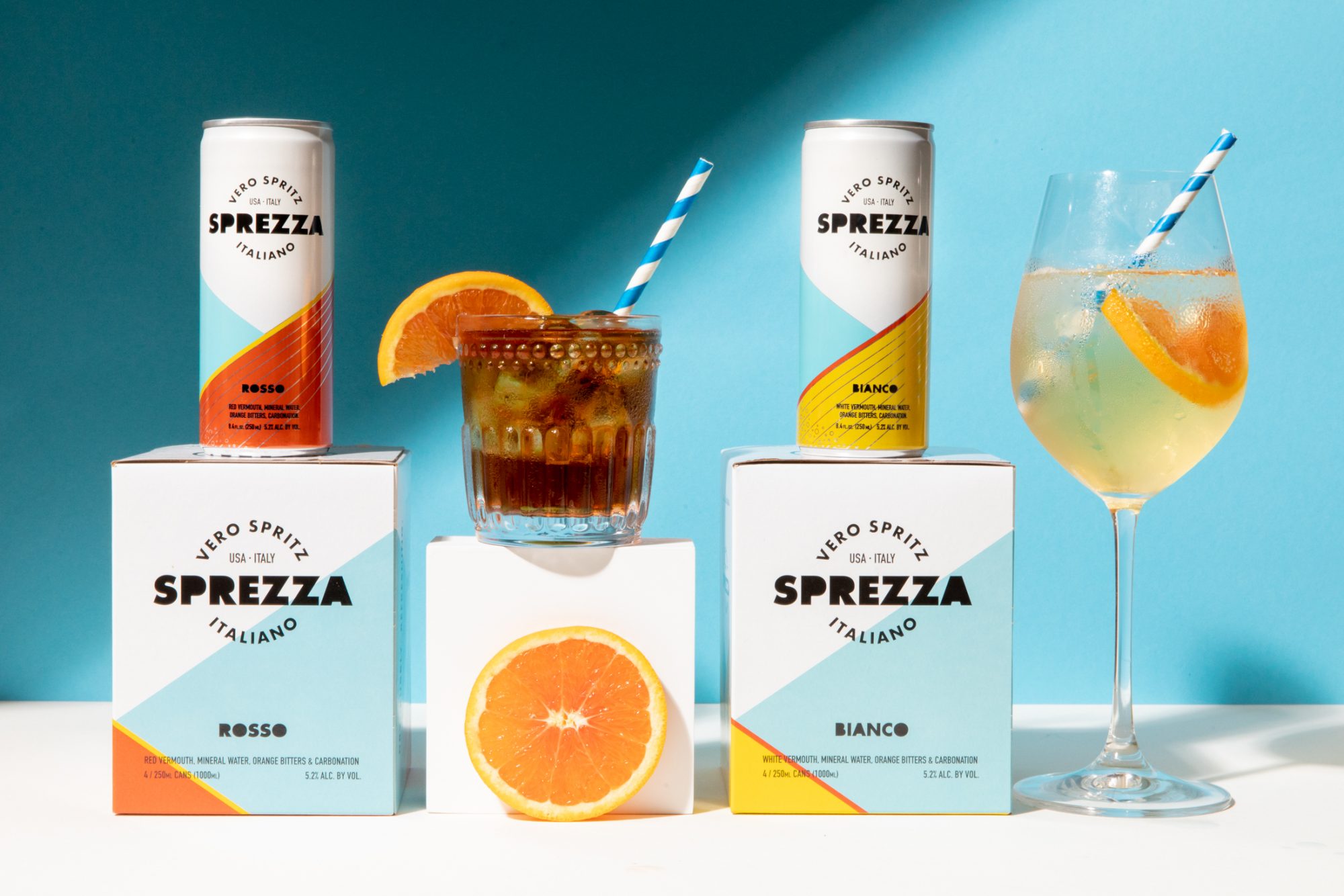 Seattle's best commercial food and hospitality photographer Two cans of Sprezza Italian spritz sit atop matching boxes, alongside a glass of dark spritz and a wine glass of pale spritz—each garnished with orange slices and striped straws. Captured by Seattles best commercial food lifestyle photographer. captured by seattle's best food and lifestyle photographer Brooke Fitts