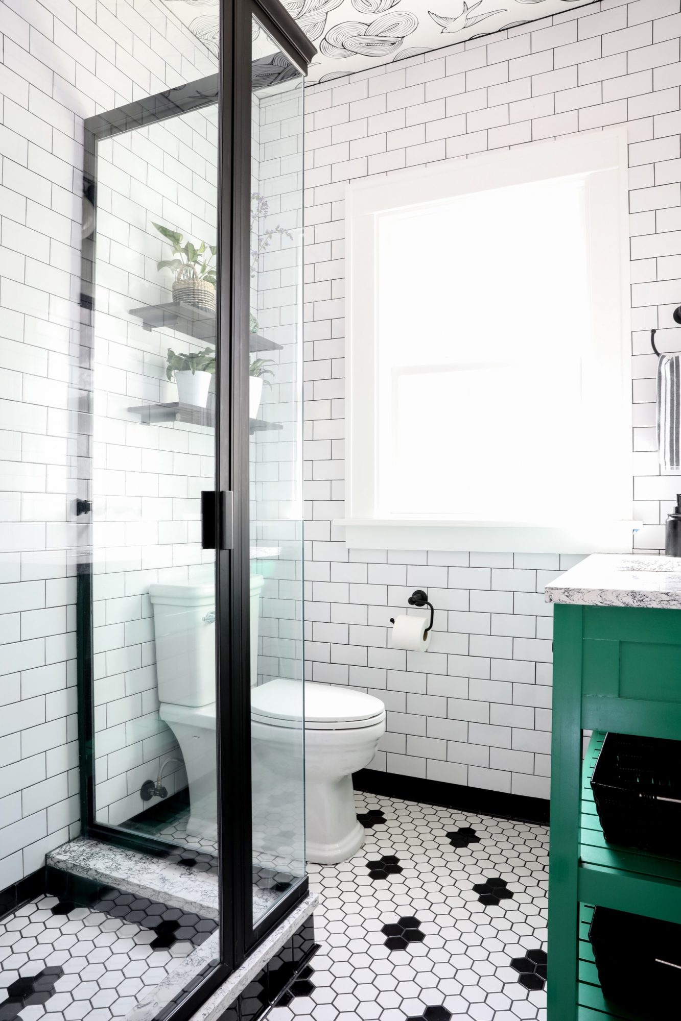 Seattle's best commercial food and hospitality photographer Bright bathroom with white subway tile walls, black grout, hexagonal black and white tile floor, and a glass shower enclosure. Captured by Seattles best commercial food lifestyle photographer, sunlight streams in over a green vanity and shelves with potted plants. captured by seattle's best food and lifestyle photographer Brooke Fitts