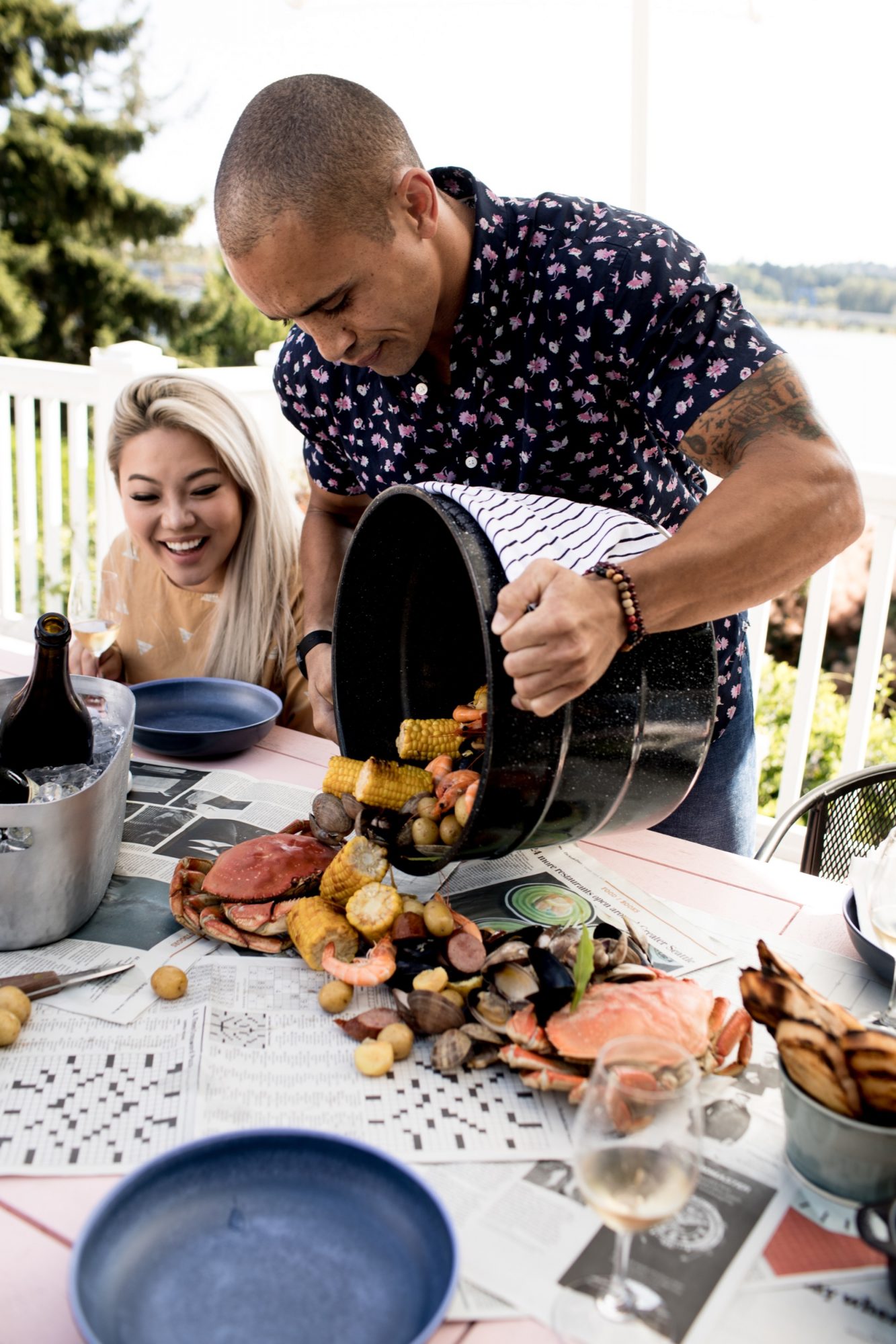Seattle's best commercial food and hospitality photographer A man pours a seafood boil with crab, corn, and potatoes onto a newspaper-covered table while a smiling woman watches. Captured outdoors with plates and wine, this moment showcases Seattles best commercial food lifestyle photographer. captured by seattle's best food and lifestyle photographer Brooke Fitts