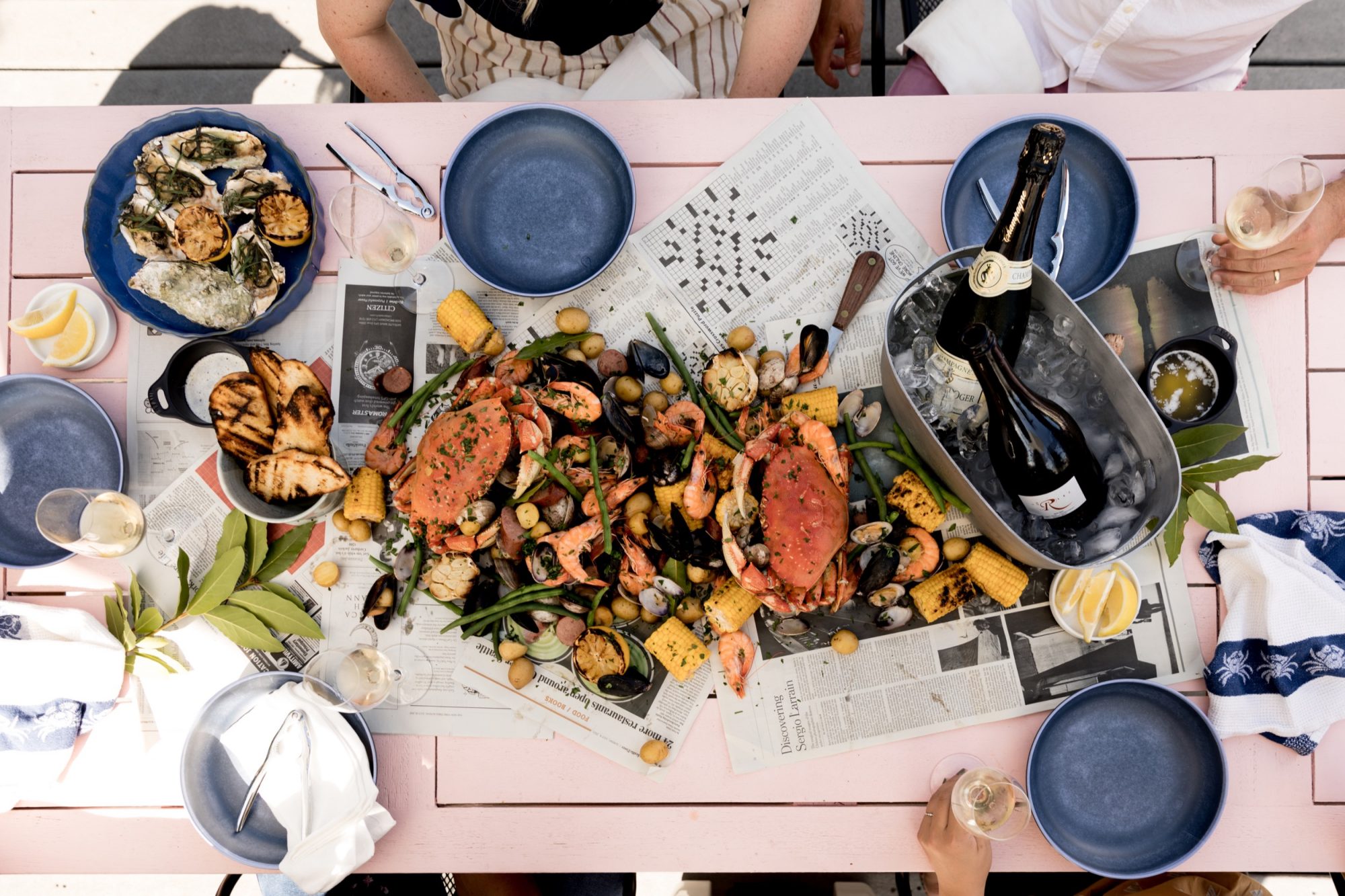 Seattle's best commercial food and hospitality photographer A pink table set with plates and glasses surrounds a seafood boil spread on newspaper, shot by Seattles best commercial food lifestyle photographer. Crab, shrimp, mussels, grilled veggies, and more tempt beside a champagne bottle chilling in ice. captured by seattle's best food and lifestyle photographer Brooke Fitts
