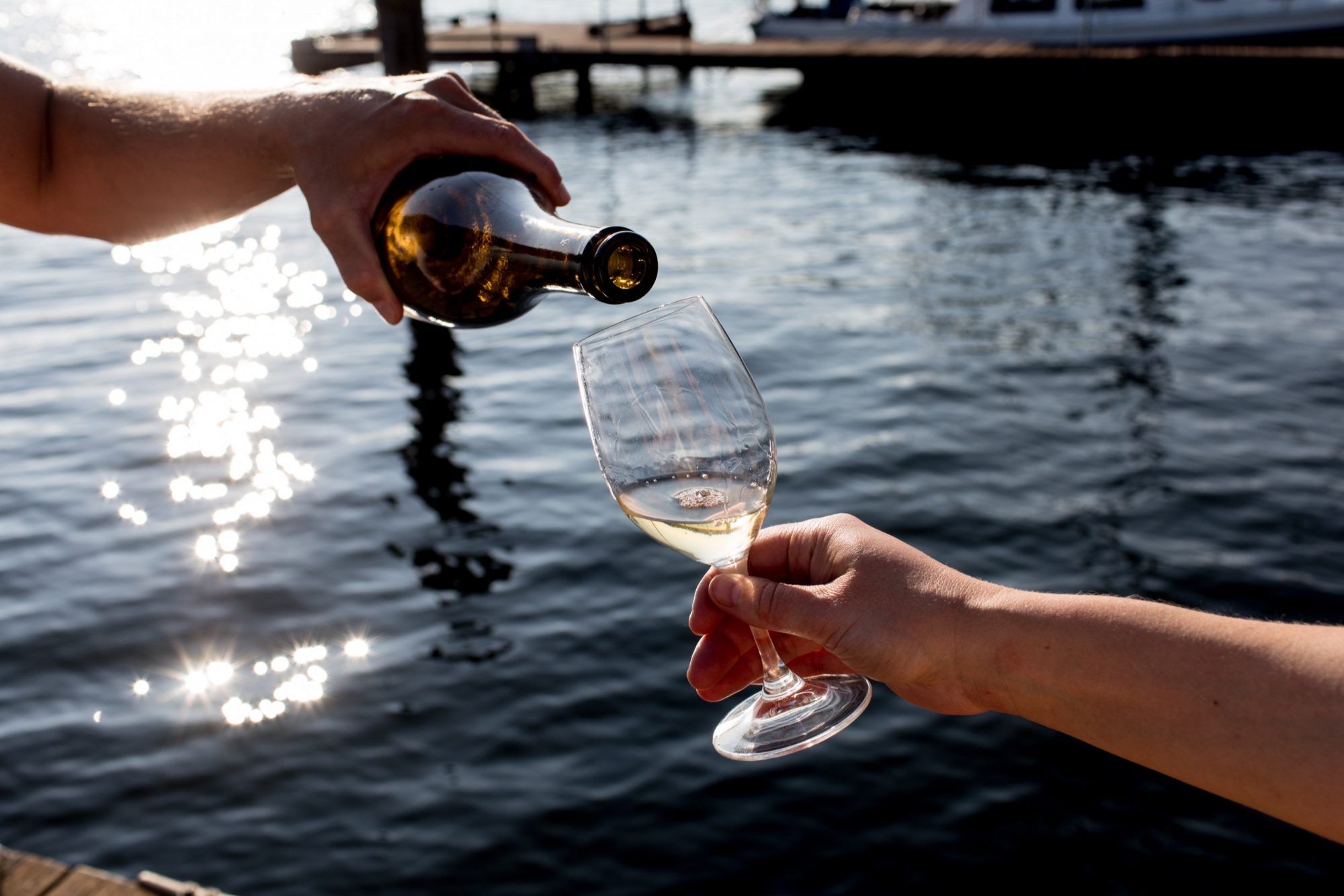 Seattle's best commercial food and hospitality photographer A hand pours white wine from a bottle into a glass held by another, with sunlight reflecting on the water and boats in the background—captured by Seattles best commercial food lifestyle photographer. captured by seattle's best food and lifestyle photographer Brooke Fitts