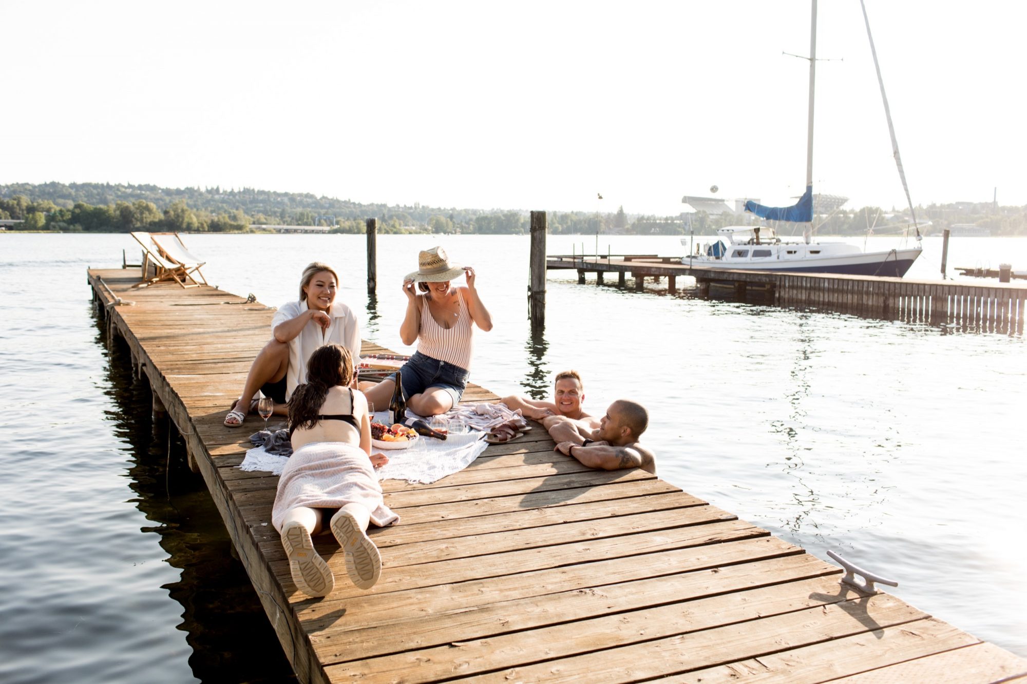 Seattle's best commercial food and hospitality photographer Five people relax on a wooden dock by the water, snacks on a towel beside them. Captured by Seattles best commercial food lifestyle photographer, this scene features friends lounging as a sailboat and trees fill the scenic background. captured by seattle's best food and lifestyle photographer Brooke Fitts