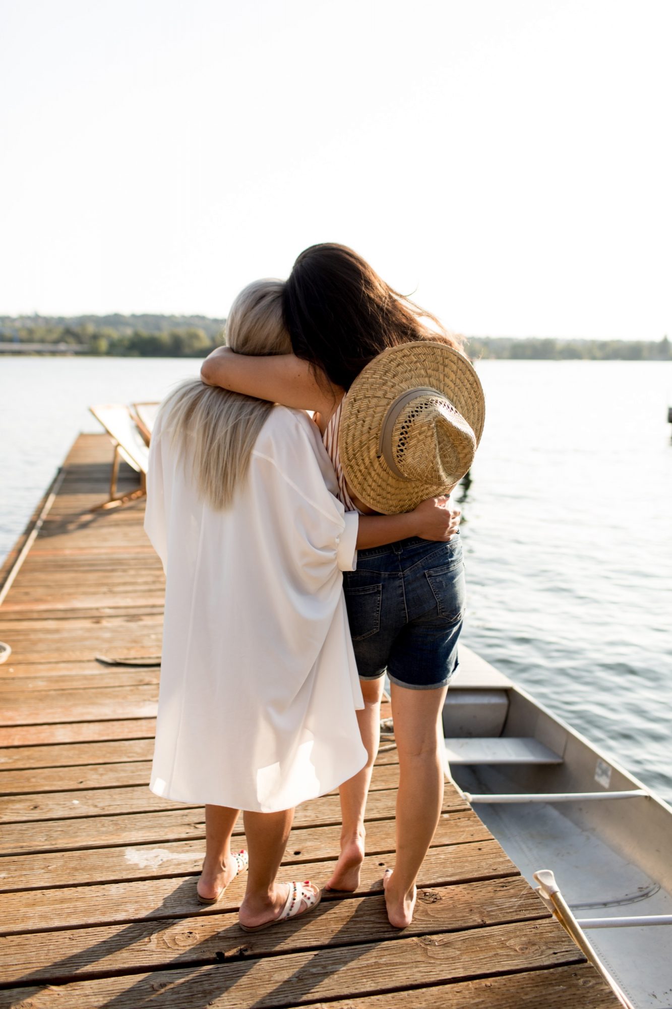 Seattle's best commercial food and hospitality photographer Two women stand arm in arm on a wooden dock by a lake, one holding a straw hat. Captured by Seattles best commercial food lifestyle photographer, both enjoy a peaceful moment together, facing the water with their backs to the camera. captured by seattle's best food and lifestyle photographer Brooke Fitts