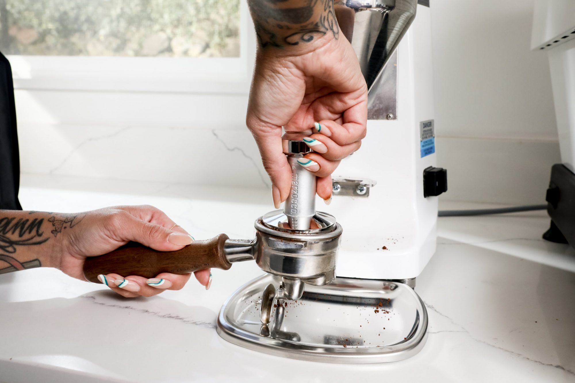 Seattle's best commercial food and hospitality photographer A person with tattoos and painted nails tamping coffee grounds in a portafilter, preparing it for an espresso machine on a white countertop—captured by Seattles best commercial food lifestyle photographer. captured by seattle's best food and lifestyle photographer Brooke Fitts