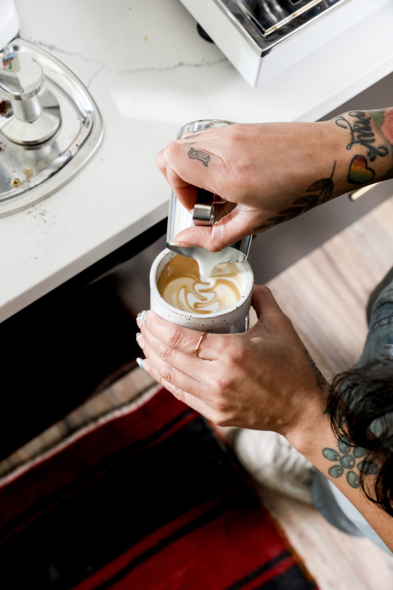 Seattle's best commercial food and hospitality photographer A person with tattoos pours steamed milk into coffee, creating latte art. Their hands hold the cup and pitcher above a white countertop with coffee equipment nearby, captured by Seattles best commercial food lifestyle photographer. captured by seattle's best food and lifestyle photographer Brooke Fitts