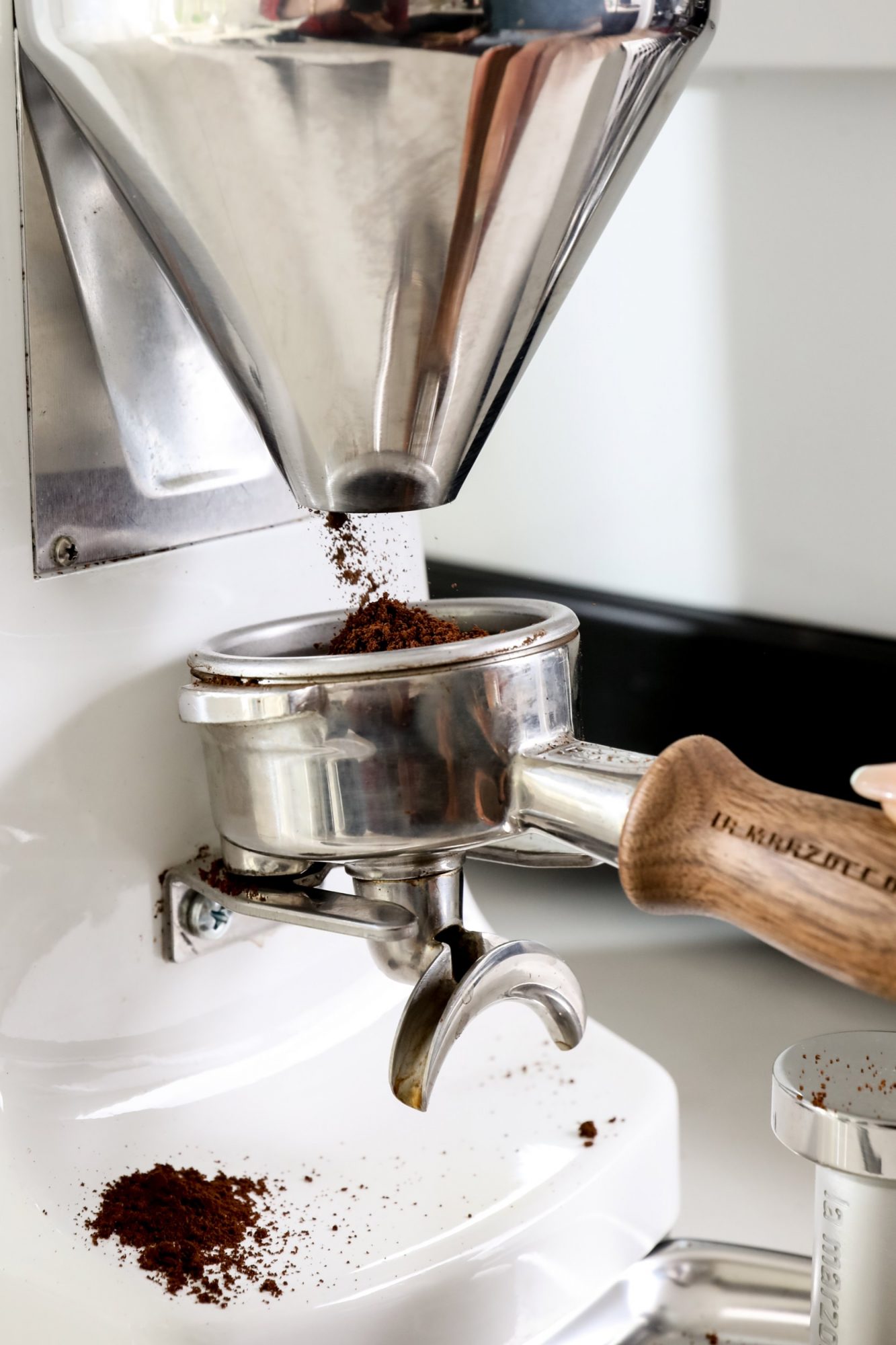 Seattle's best commercial food and hospitality photographer A close-up by Seattles best commercial food lifestyle photographer shows ground coffee dispensing from a grinder into a portafilter with a wooden handle, scattered coffee grounds dotting the white counter below. captured by seattle's best food and lifestyle photographer Brooke Fitts