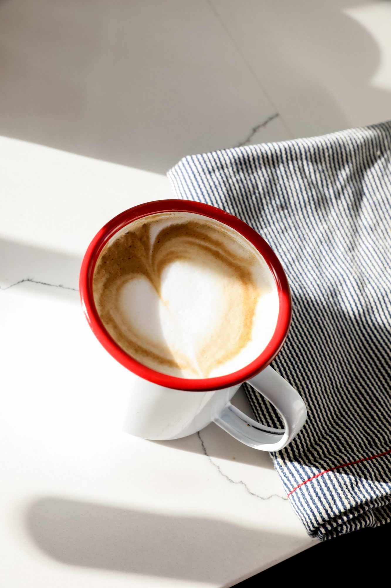 Seattle's best commercial food and hospitality photographer A white mug with a red rim holds a cappuccino with foam art, styled by Seattles best commercial food lifestyle photographer, placed on a white countertop next to a blue-and-white striped cloth in bright sunlight. captured by seattle's best food and lifestyle photographer Brooke Fitts