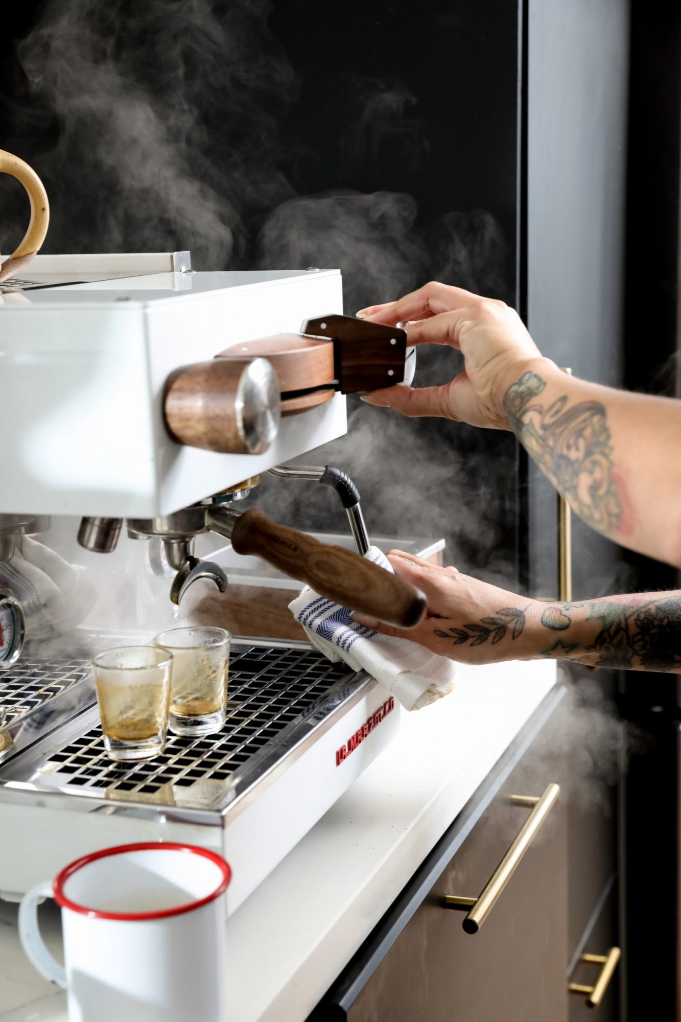 Seattle's best commercial food and hospitality photographer A tattooed barista operates a steaming espresso machine, pulling wooden handles as two small glasses fill with espresso. Captured by Seattles best commercial food lifestyle photographer, a white mug with a red rim sits nearby on the counter. captured by seattle's best food and lifestyle photographer Brooke Fitts