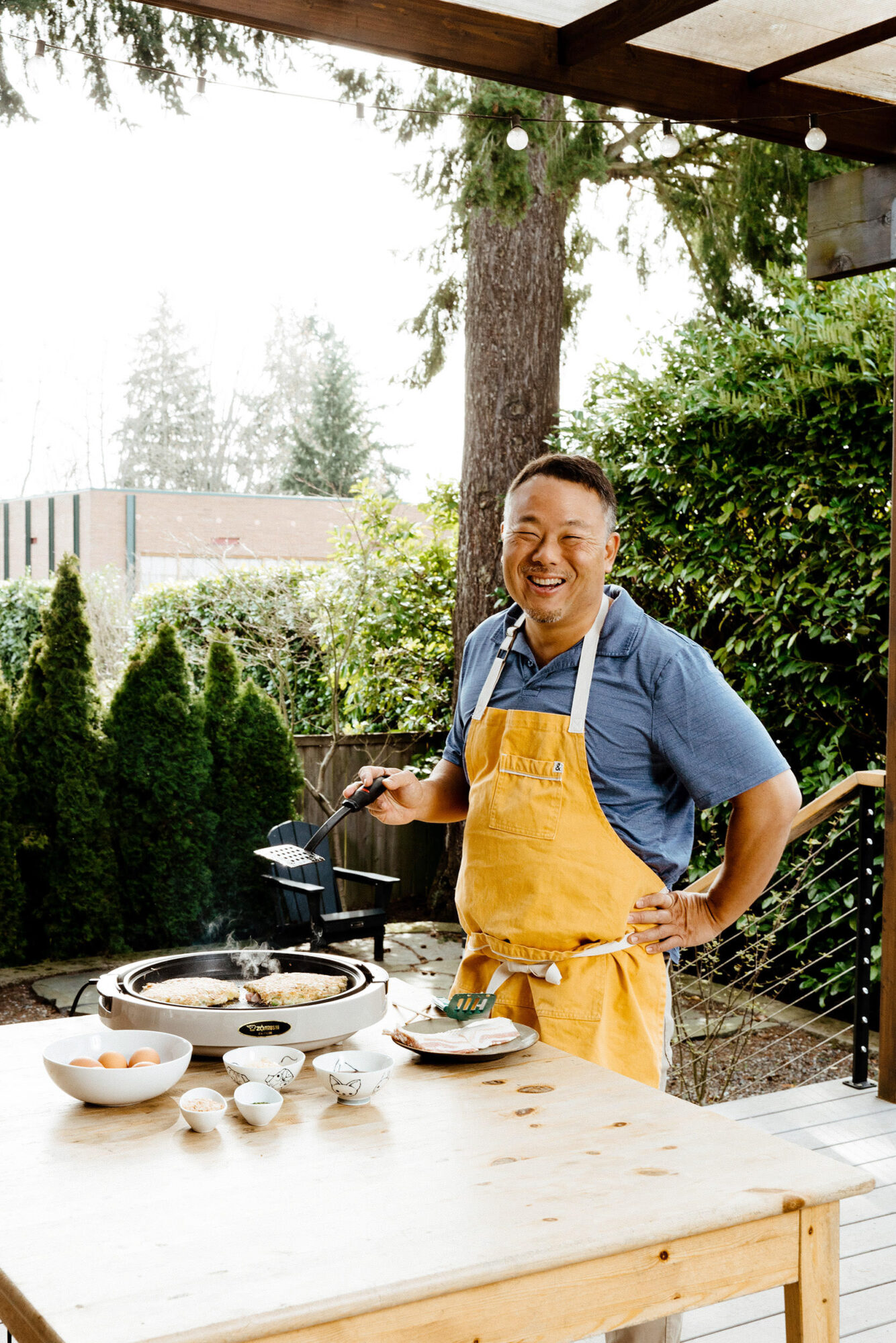 Seattle's best commercial food and hospitality photographer A man wearing a yellow apron stands outdoors by a grill, smiling while cooking. Captured by Seattles best commercial food lifestyle photographer, bowls of ingredients sit on a wooden table amid lush greenery and trees in the background. captured by seattle's best food and lifestyle photographer Brooke Fitts