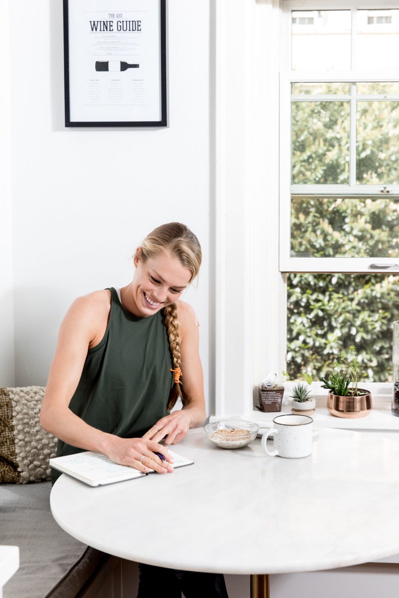 Seattle's best commercial food and hospitality photographer A woman with a blonde braid, wearing a sleeveless dark green top, smiles while writing in a notebook at a round white table—a scene captured by Seattles best commercial food lifestyle photographer. A mug, bowl, and potted plants bask near a sunny window. captured by seattle's best food and lifestyle photographer Brooke Fitts