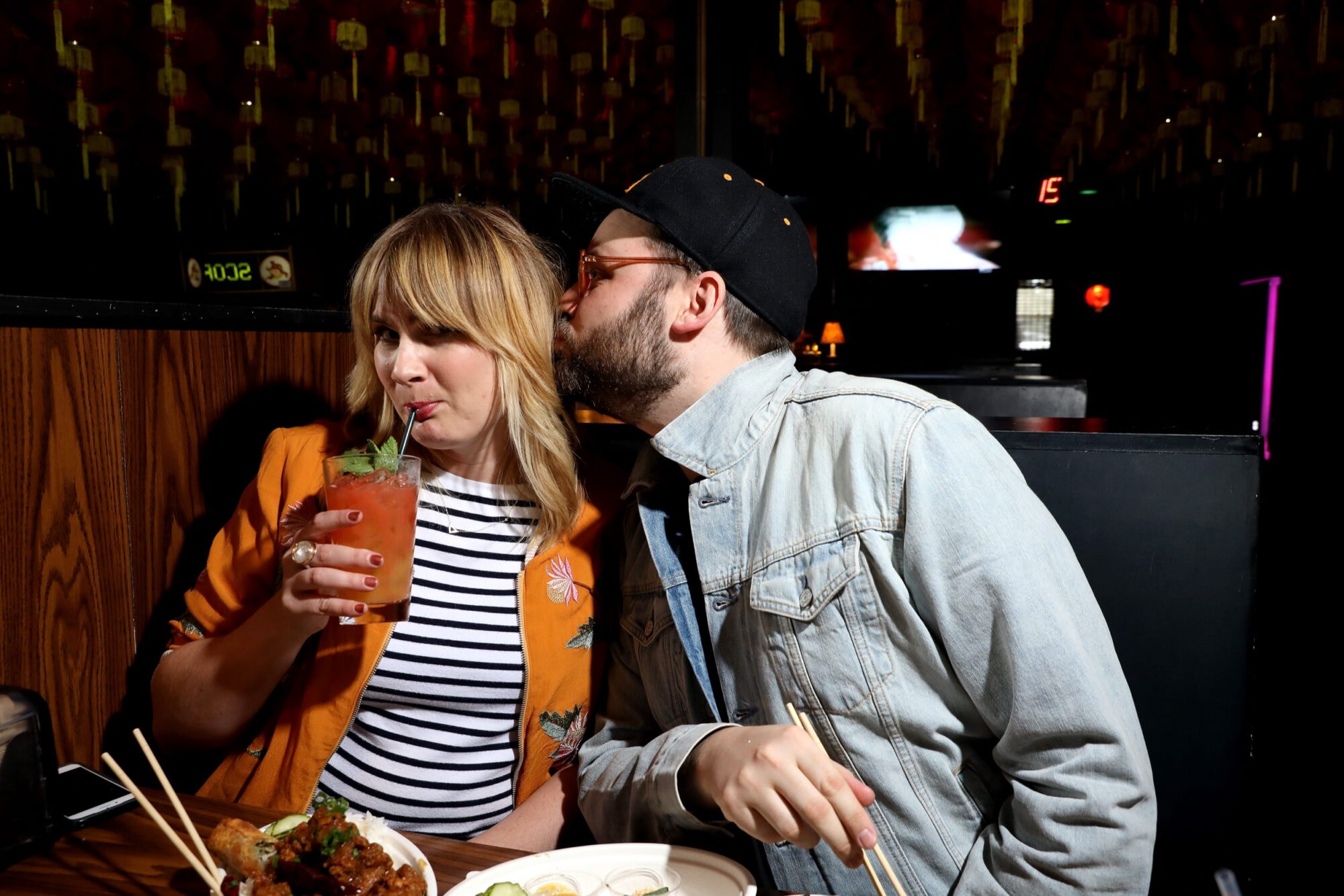 Seattle's best commercial food and hospitality photographer A man in a denim jacket and cap kisses a woman on the cheek as she drinks a red cocktail at a restaurant table, captured by Seattles best commercial food lifestyle photographer. The woman smiles in her striped shirt and orange jacket. captured by seattle's best food and lifestyle photographer Brooke Fitts