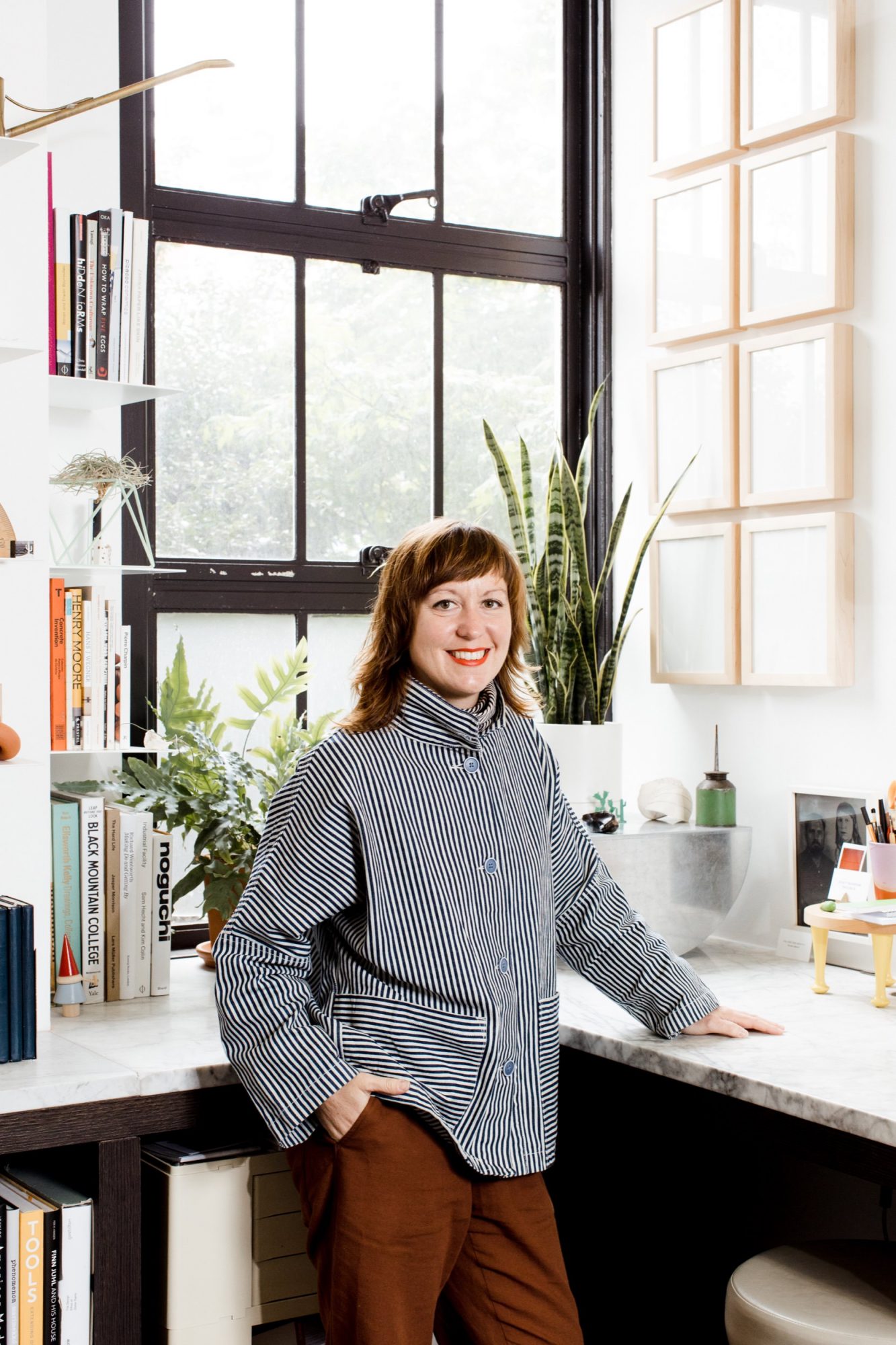 Seattle's best commercial food and hospitality photographer A person with shoulder-length hair wearing a striped shirt stands and smiles in a bright, modern office—captured by Seattles best commercial food lifestyle photographer—surrounded by large windows, plants, books, and framed art. captured by seattle's best food and lifestyle photographer Brooke Fitts