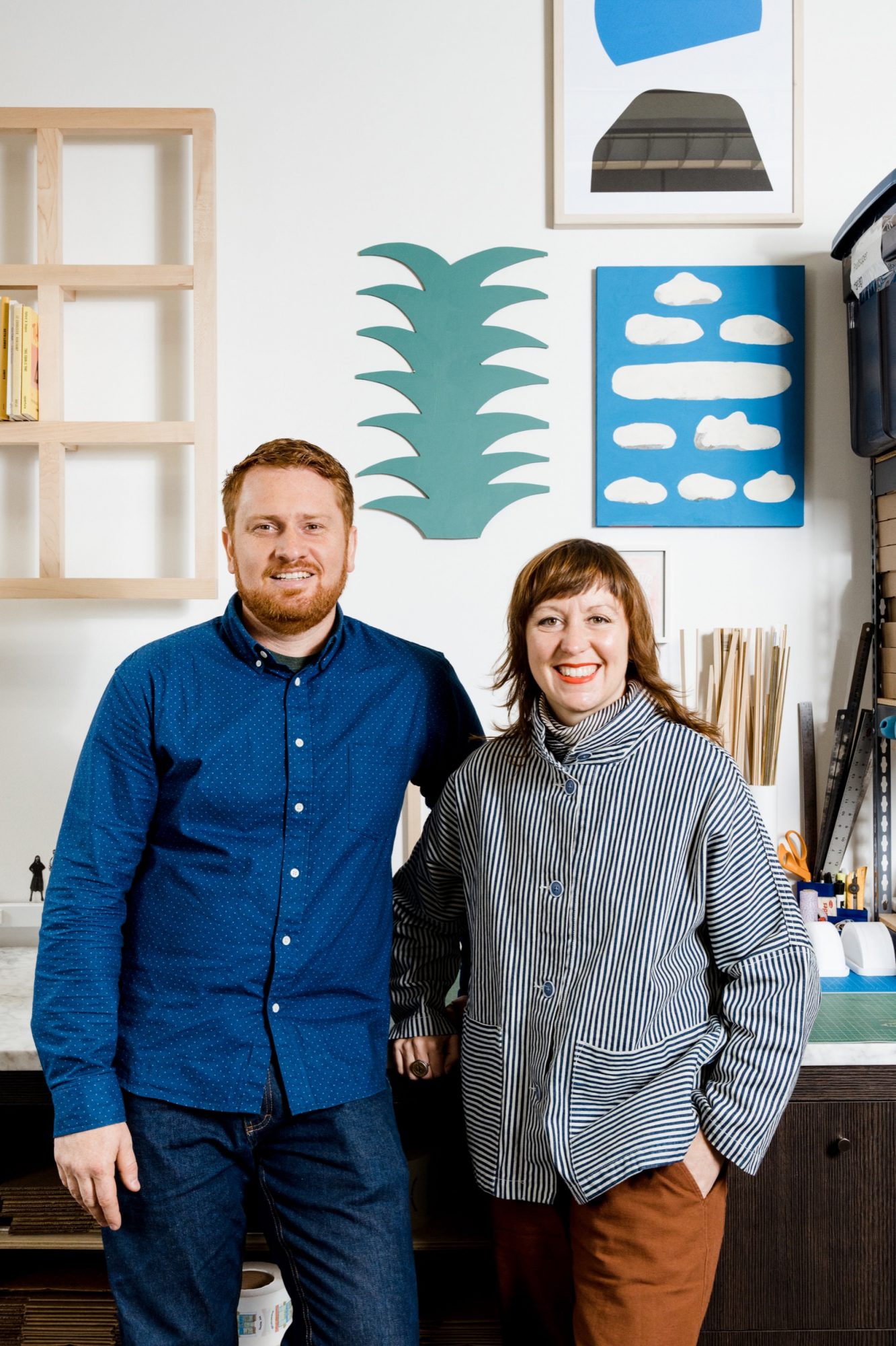 Seattle's best commercial food and hospitality photographer A man and woman stand smiling in a creative studio with abstract art and craft supplies in the background, captured by Seattles best commercial food lifestyle photographer. The man wears a blue shirt and jeans; the woman wears a striped jacket and brown pants. captured by seattle's best food and lifestyle photographer Brooke Fitts
