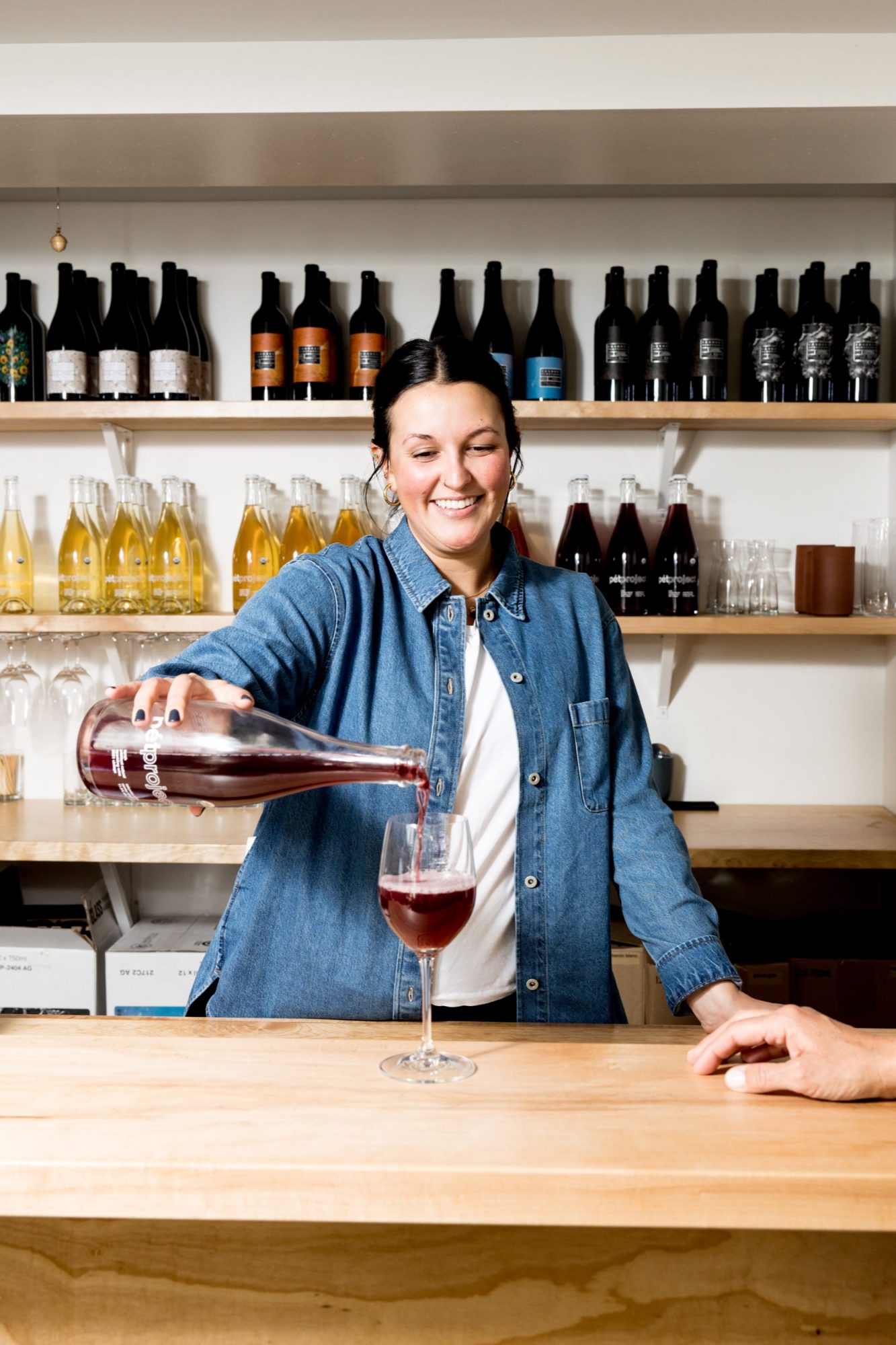 Seattle's best commercial food and hospitality photographer A smiling woman in a denim shirt pours red wine into a glass at a wooden counter, captured by Seattles best commercial food lifestyle photographer. Shelves of wine bottles and glasses fill the background, with a hand resting on the counter in front. captured by seattle's best food and lifestyle photographer Brooke Fitts