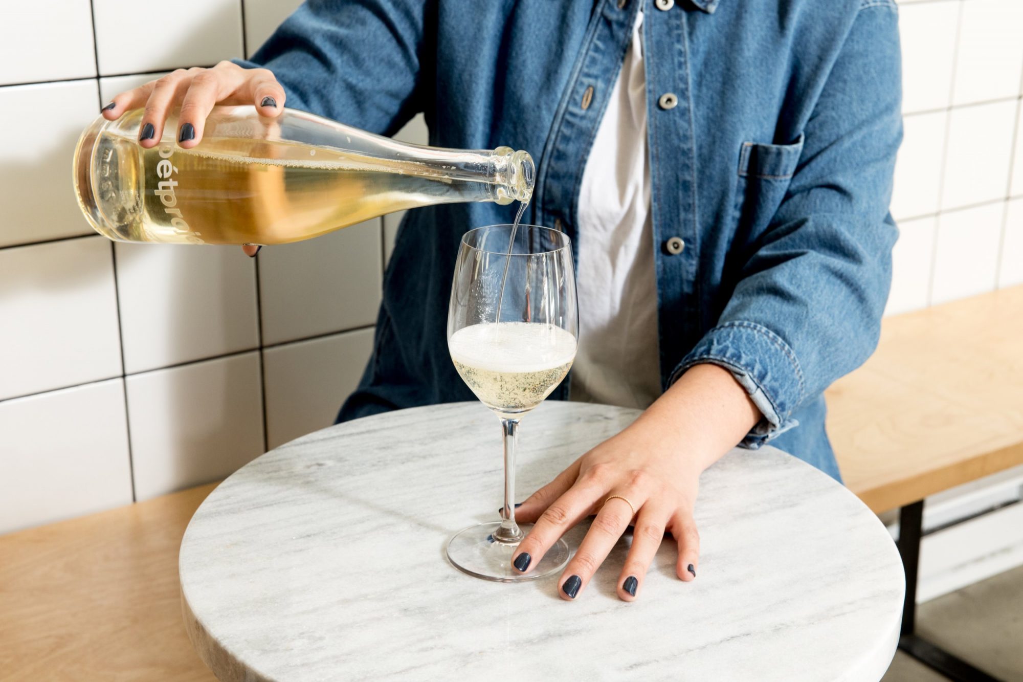 Seattle's best commercial food and hospitality photographer A person in a denim shirt pours sparkling wine from a bottle into a glass on a round marble table—captured beautifully by Seattles best commercial food lifestyle photographer, with white tiled walls and a wooden bench in the background. captured by seattle's best food and lifestyle photographer Brooke Fitts