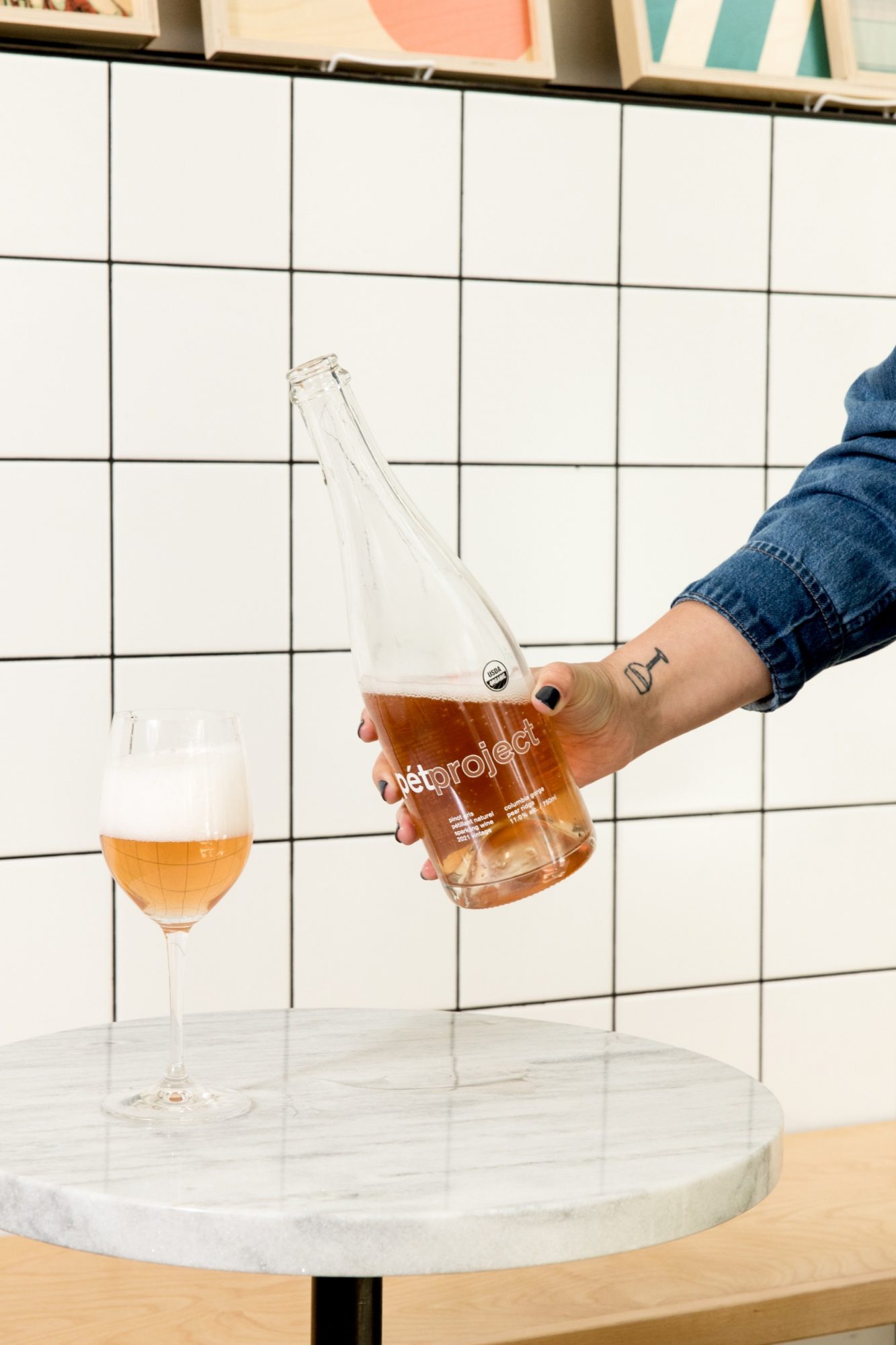 Seattle's best commercial food and hospitality photographer Seattles best commercial food lifestyle photographer captures a hand pouring orange-colored Outproject drinks into a foamy stemmed glass, set on a small round marble table against a white tiled wall. captured by seattle's best food and lifestyle photographer Brooke Fitts