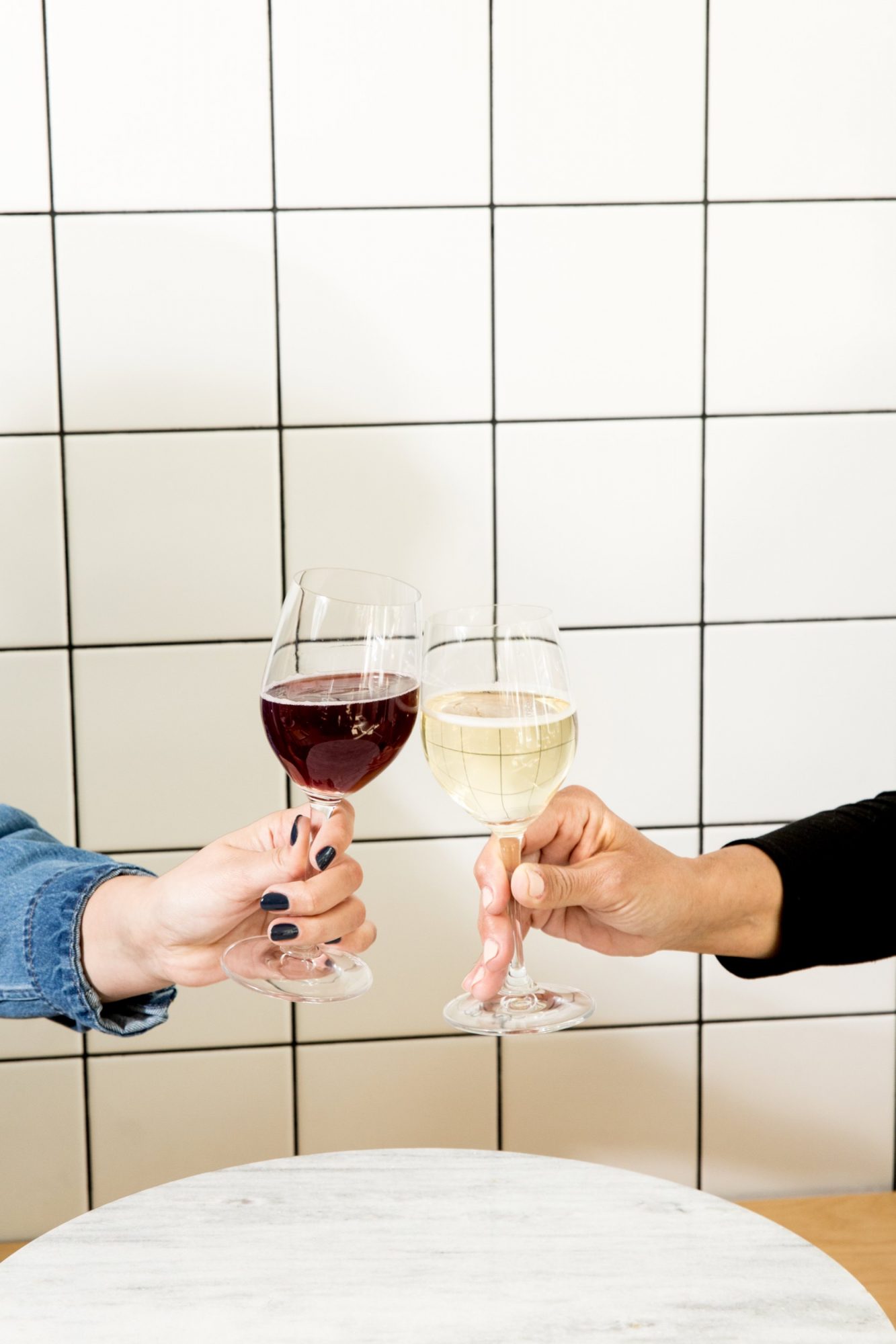 Seattle's best commercial food and hospitality photographer Two people clink glasses of red and white wine over a round white table, with a white tiled wall in the background—a vibrant moment captured by Seattles best commercial food lifestyle photographer. One person wears blue, the other black. captured by seattle's best food and lifestyle photographer Brooke Fitts