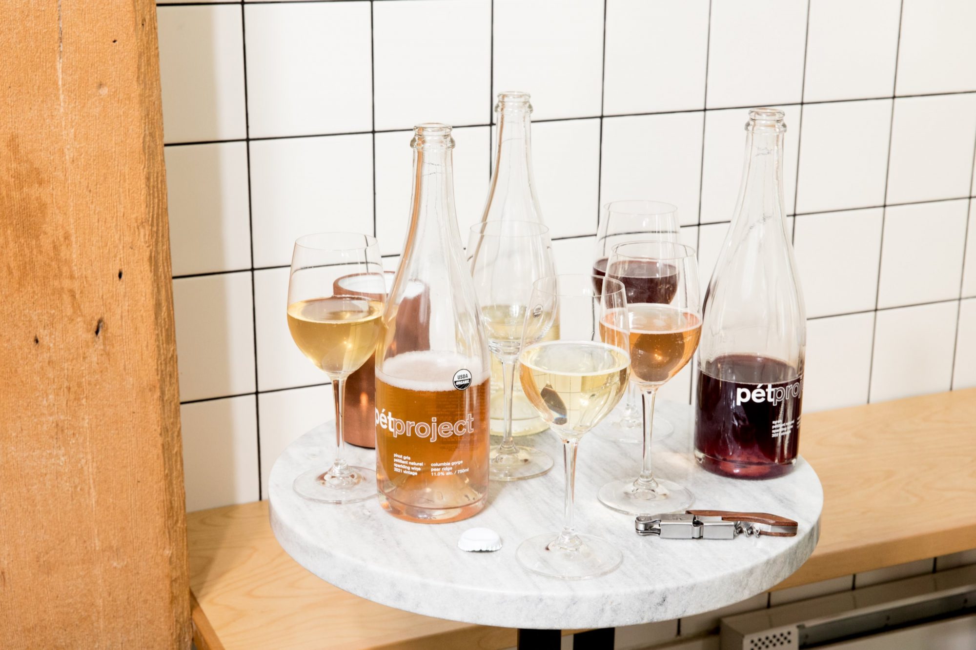 Seattle's best commercial food and hospitality photographer A round marble table set by Seattles best commercial food lifestyle photographer showcases wine glasses filled with different wines, three labeled bottles, a corkscrew, and cork, against white tiled and wooden walls. captured by seattle's best food and lifestyle photographer Brooke Fitts