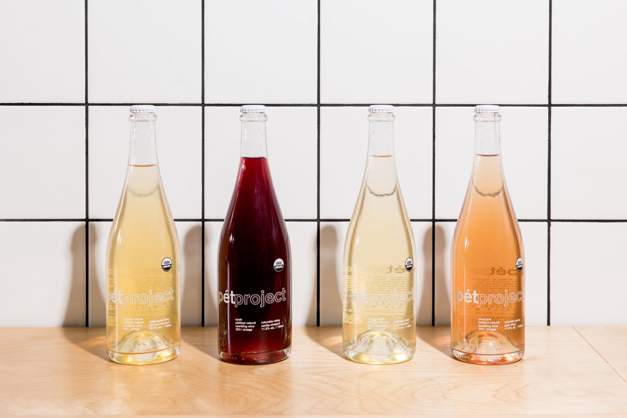 Seattle's best commercial food and hospitality photographer Four bottles of pét-nat wine in clear glass, each a different hue, stand in a row on wood against white tiles—captured by Seattles best commercial food lifestyle photographer. captured by seattle's best food and lifestyle photographer Brooke Fitts